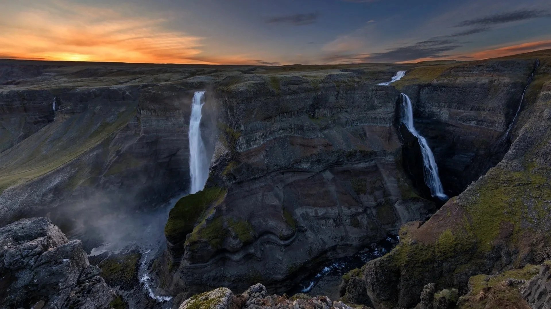 Mehrere Wasserfälle fließen durch eine zerklüftete, grüne Felslandschaft bei Sonnenuntergang, mit einem bewölkten Himmel im Hintergrund.