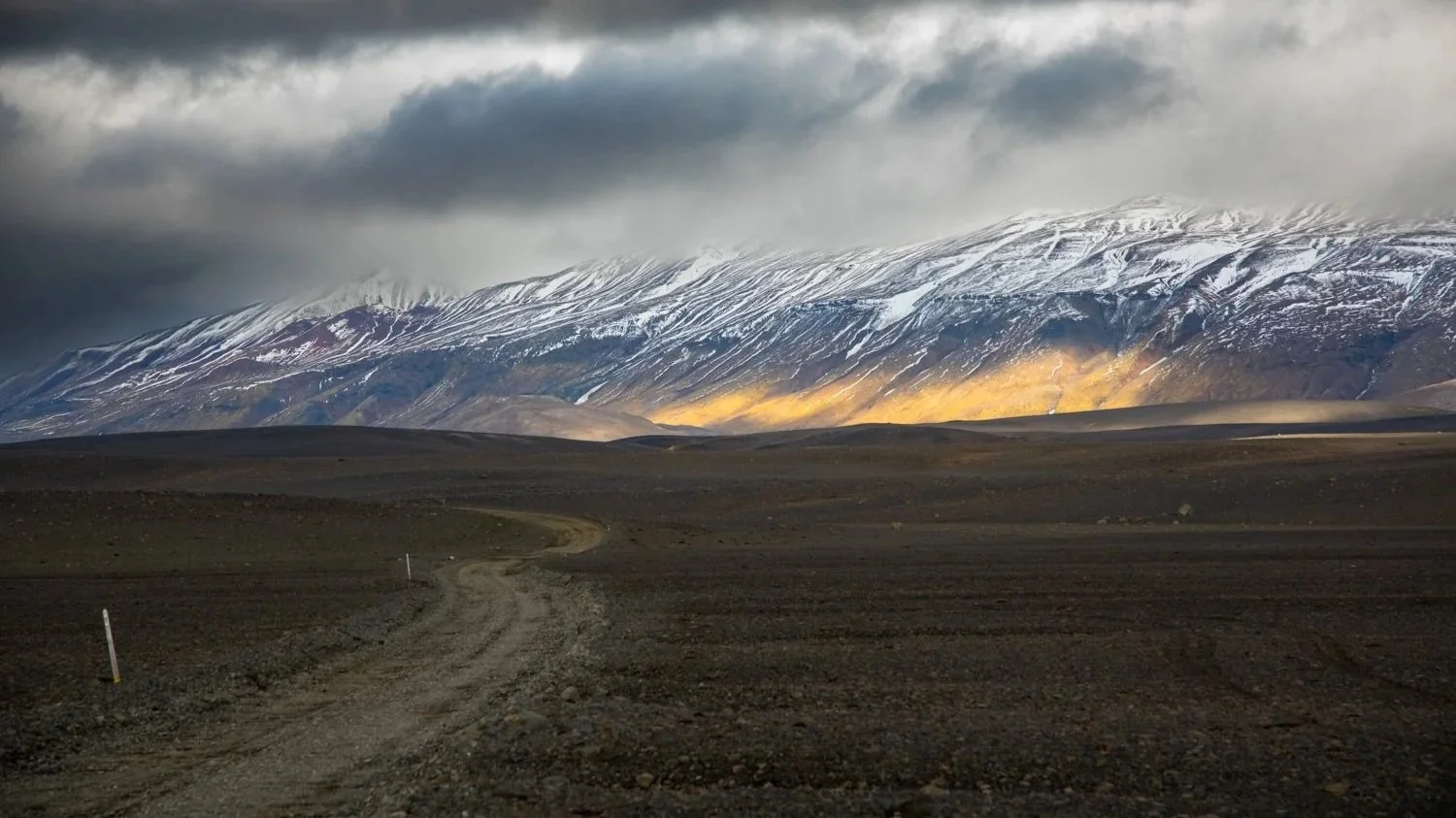 Landschaft mit einer unbefestigten Straße im Vordergrund, weite Ebenen und hohe Berge mit Schneeflächen im Hintergrund, dunkle Wolken am Himmel.