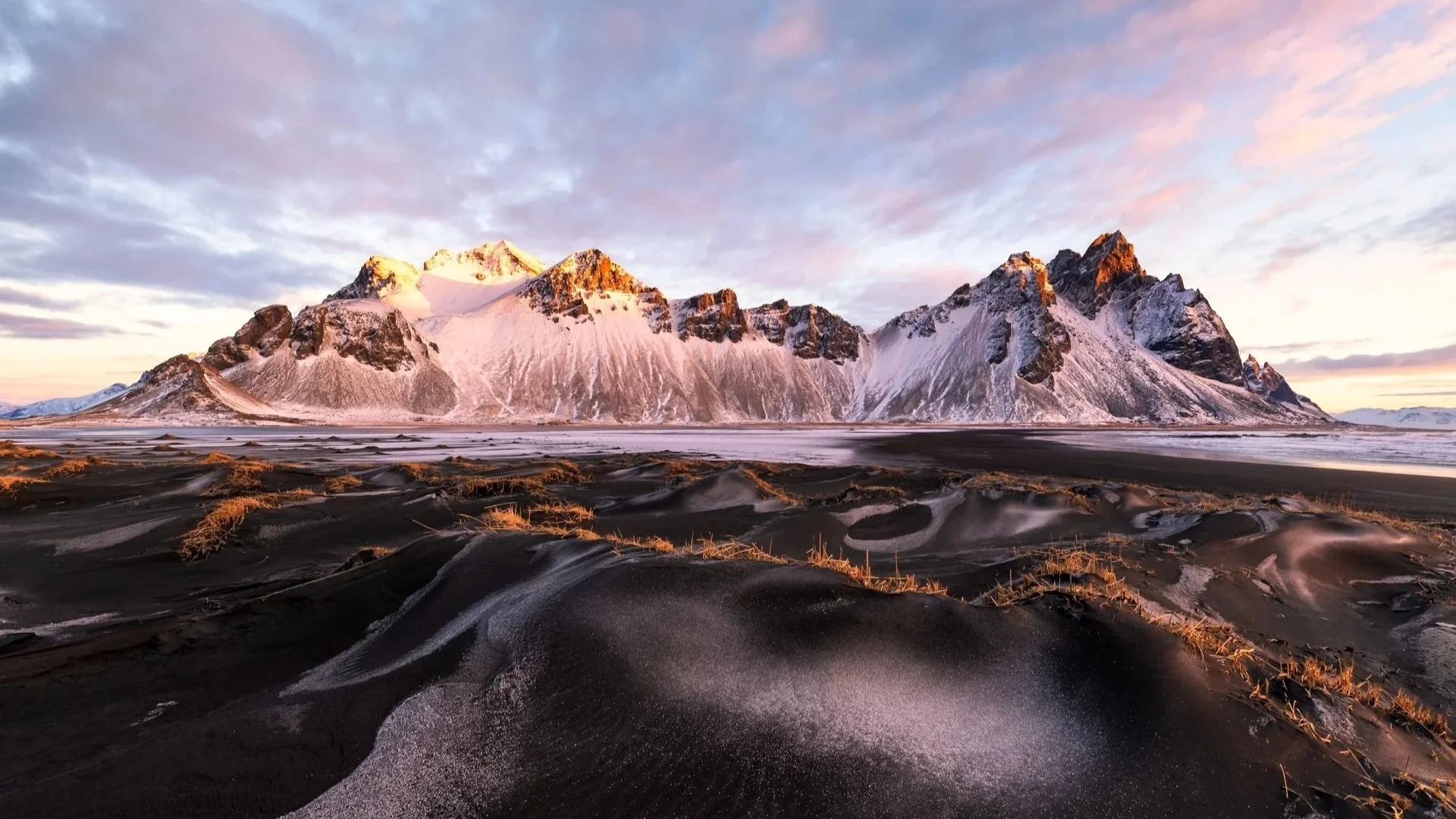 Schneebedeckte Berge bei Sonnenuntergang, dunkler Sandstrand im Vordergrund, skandinavische Landschaft
