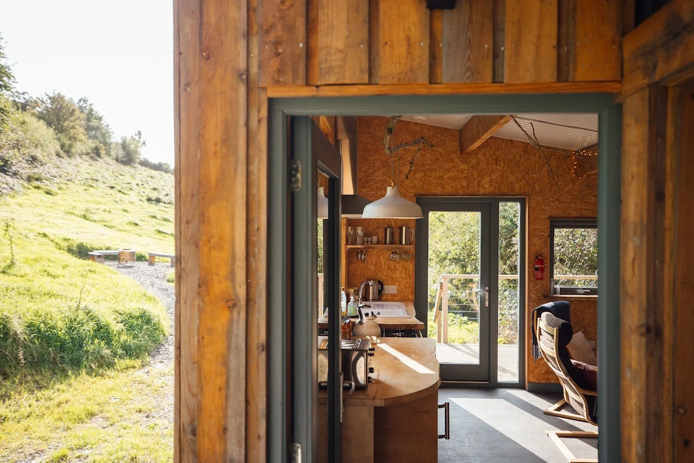 The left of the shot is outside Ty Mawr, view of the grassy hill, the right is the doorway and interior of kitchen dining area with modern white lamp hanging down, all timberwork and warm looking.