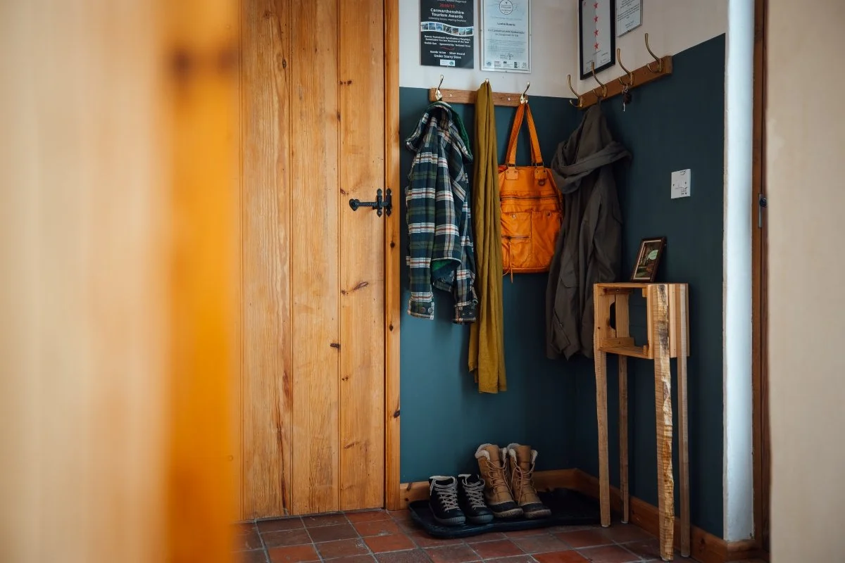 Hall detail with rustic timber door and black latch, deep blue walls and hooks with coats and walking boots below.