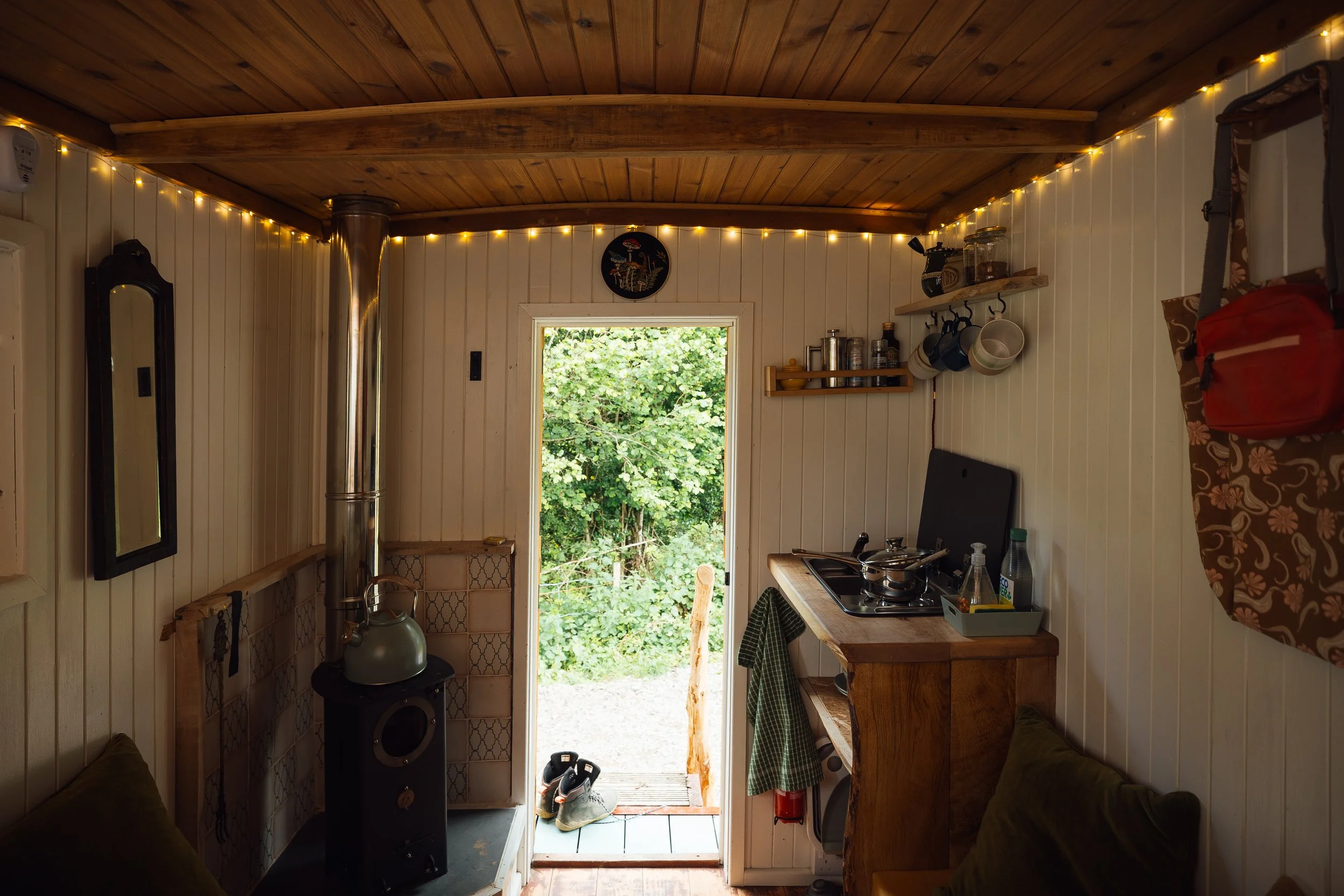 Interior of a small cozy cabin kitchen with wooden ceilings and white paneled walls. There is a door leading outside with greenery visible beyond. The room has a chimney with a kettle on it, a small stove, a shelf with mugs and jars, and other kitche