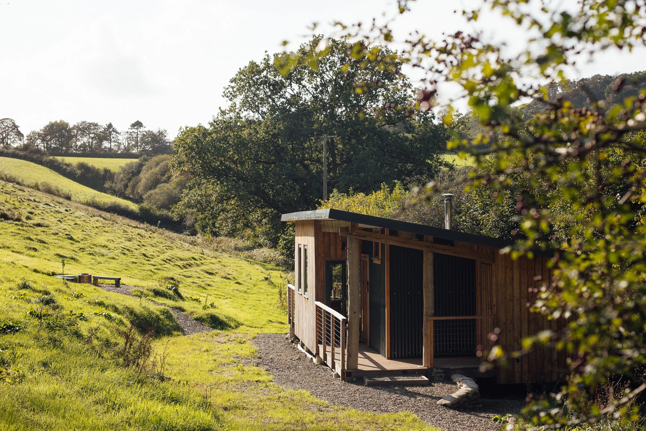 Ty Mawr in its landscape, at the base of a grassy slope, a timber building with an open railed porch, a metal chimney flu and large picture windows and doors.