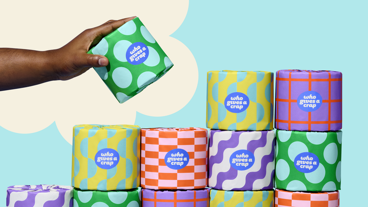 Image shows a stack of colourfully wrapped rolls of toilet paper from the brand "Who Gives A Crap"  with a hand taking a roll from the top of the stack, with a background of a blue sky with a white fluffy cloud illustration.