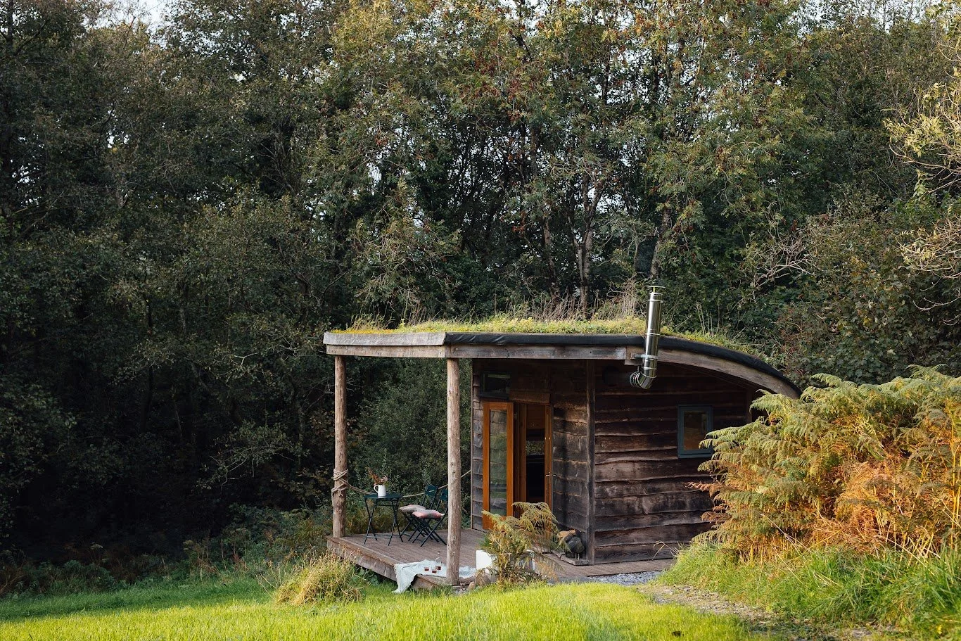 External view of Hafan Cabin, clad in dark waney edge timber with its timber porch deck with two chairs and a table.  The cabin has a green roof and is set in front of mature trees in leaf.
