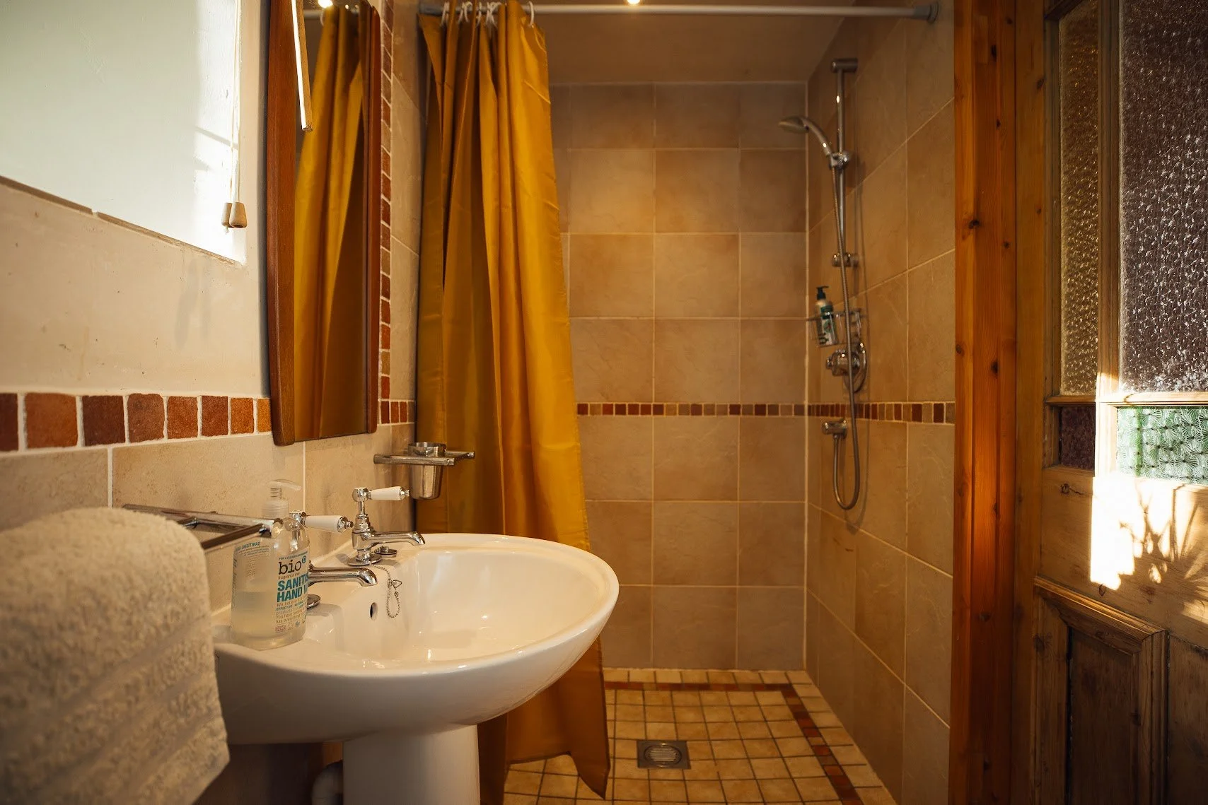 Bathroom at Llwynbwch Barn with warm light brown tiles to shower cubicle, mustard shower curtain, chrome shower, white sink to left with traditional design (accessible) taps.  Tiled floor and glimpse of window to right.