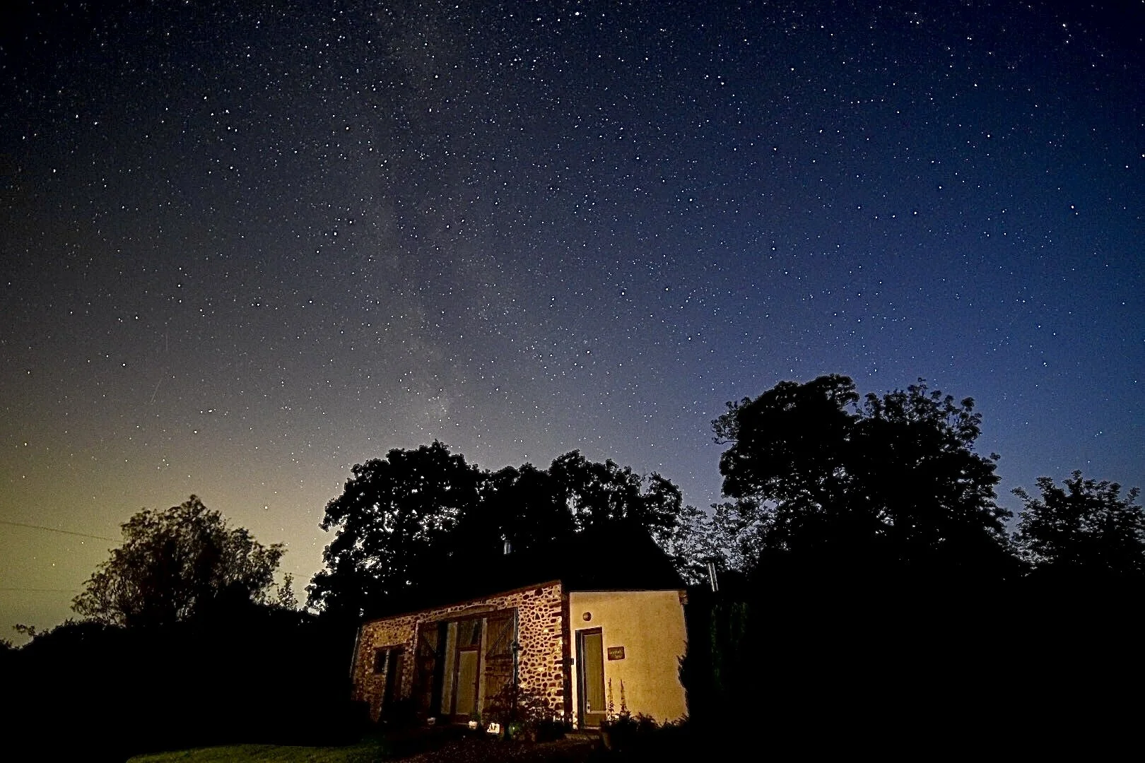 Llwynbwch Barn under dark starry skies, the stone frontage lit up and silhouetted by trees. Dark blue, orange and mid blue sky above with columns of light and stars throughout the sky.