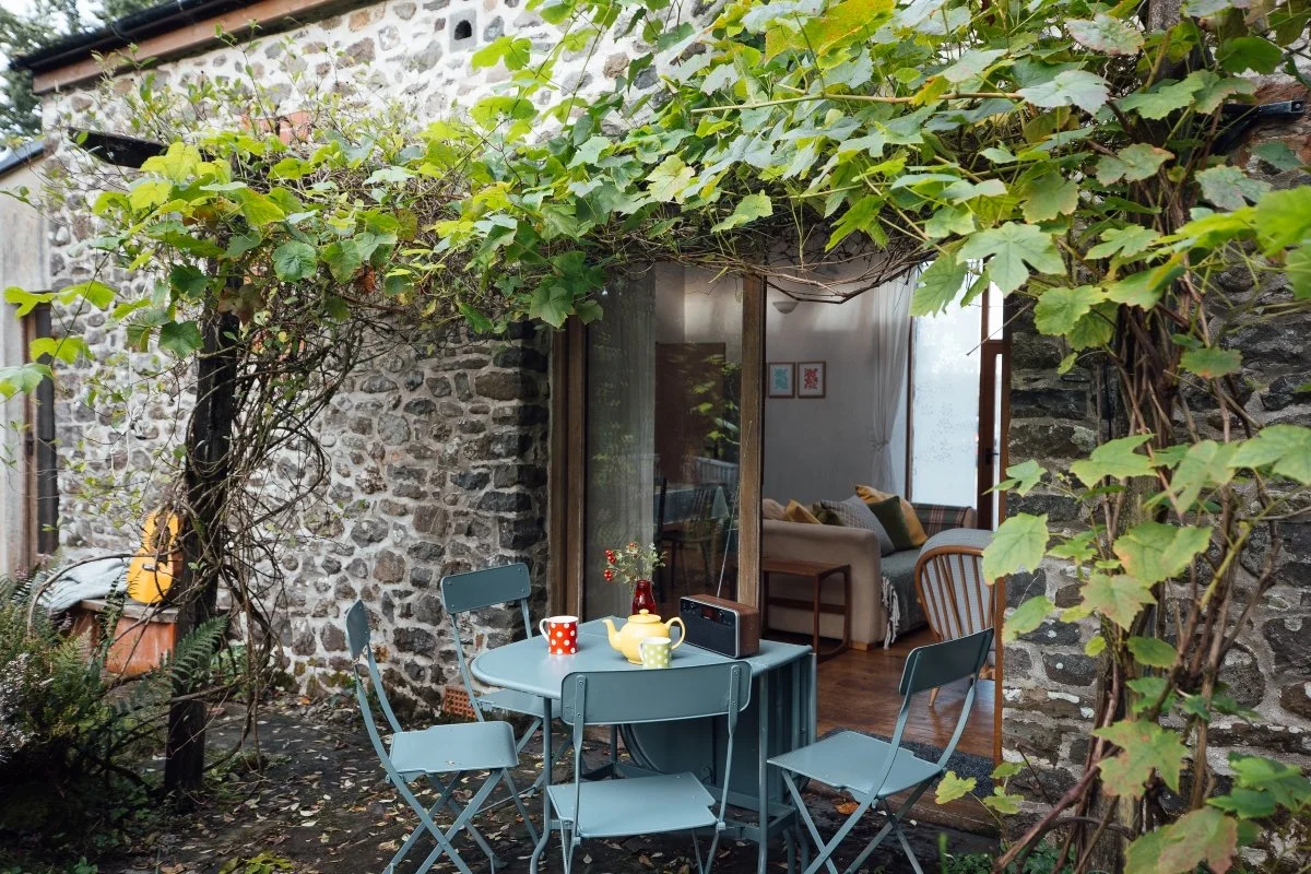 Patio dining area at Llwnbwch Barn with climbing plant up and over wooden french doors set in random rubble stone walls. Light blue metal dining table and chairs laid for breakfast with colourful china cups and teapot,