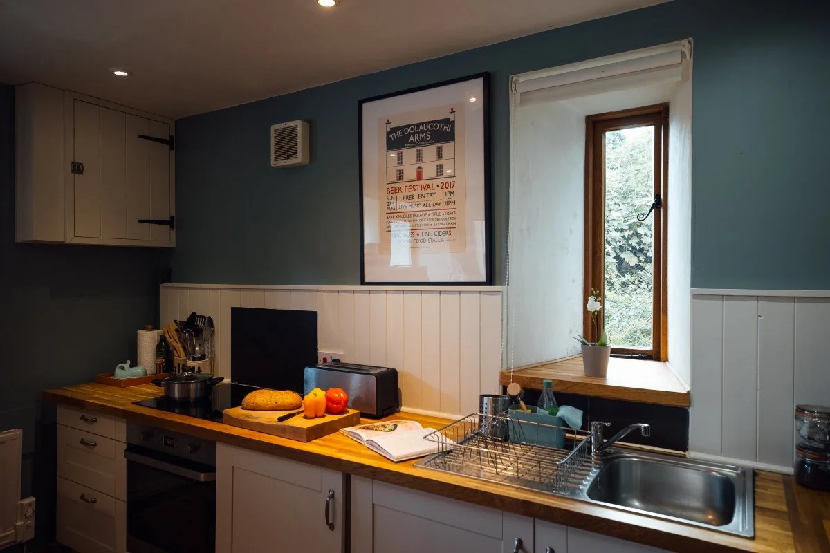 Kitchen at Llwynbwch with soft teal blue painted walls, wooden counter and white cupboards below, showing sink, chopping board with fruit, black cooker (oven & hob) and window onto garden beyond.