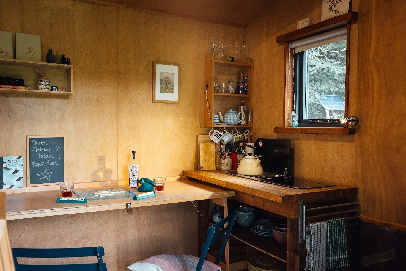 Inside Hafan Cabin showing the kitchen and table area in the corner of the cabin. Wooden walls and kitchen crockery including a kettle, drinks and glasses.