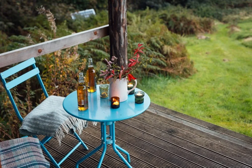Detail of the blue metal table and chairs, with blankets, at Derwen Cabin, table laid with wildflowers and maple leaves, with two bottles of beer and tea lights in coloured glass holders. The post and rail of the deck with green setting beyond.