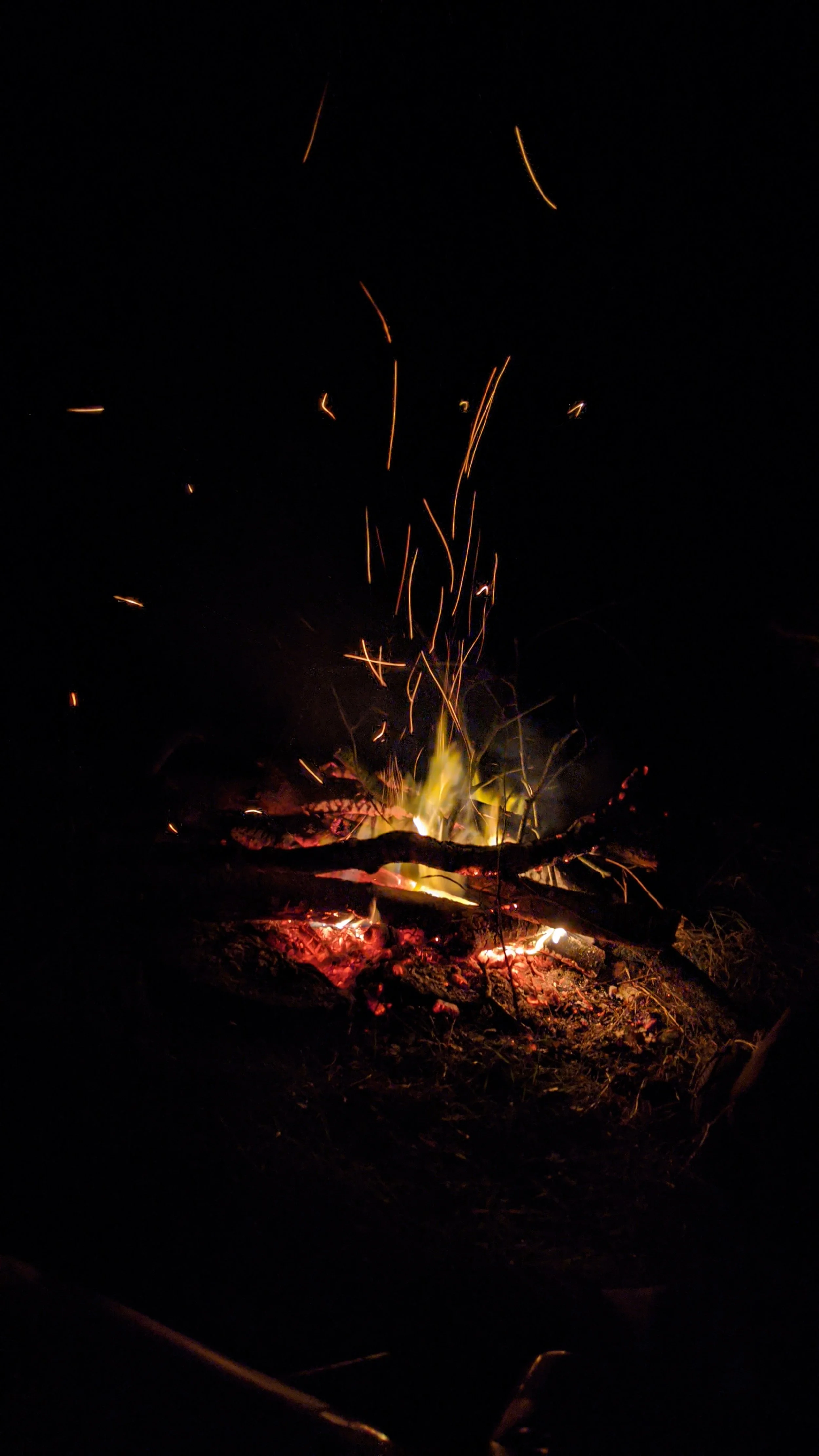 A campfire at night - the whole picture is dark apart from the flames rising up through the logs and long embers of light streaming up into the air.