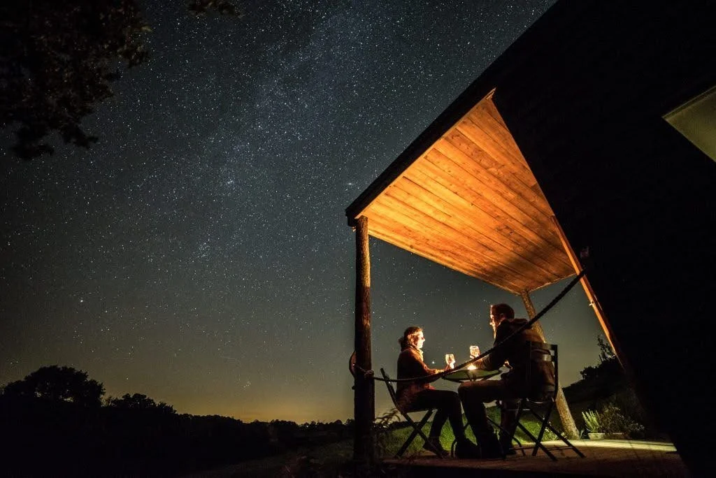 Stargazing on the deck at Derwen Cabin. Two seated figures enjoying drinks at a table under star studded dark skies, the land below in dark silhouette. Warm orange light on their faces and lighting up the inside of the cabin deck overhang.