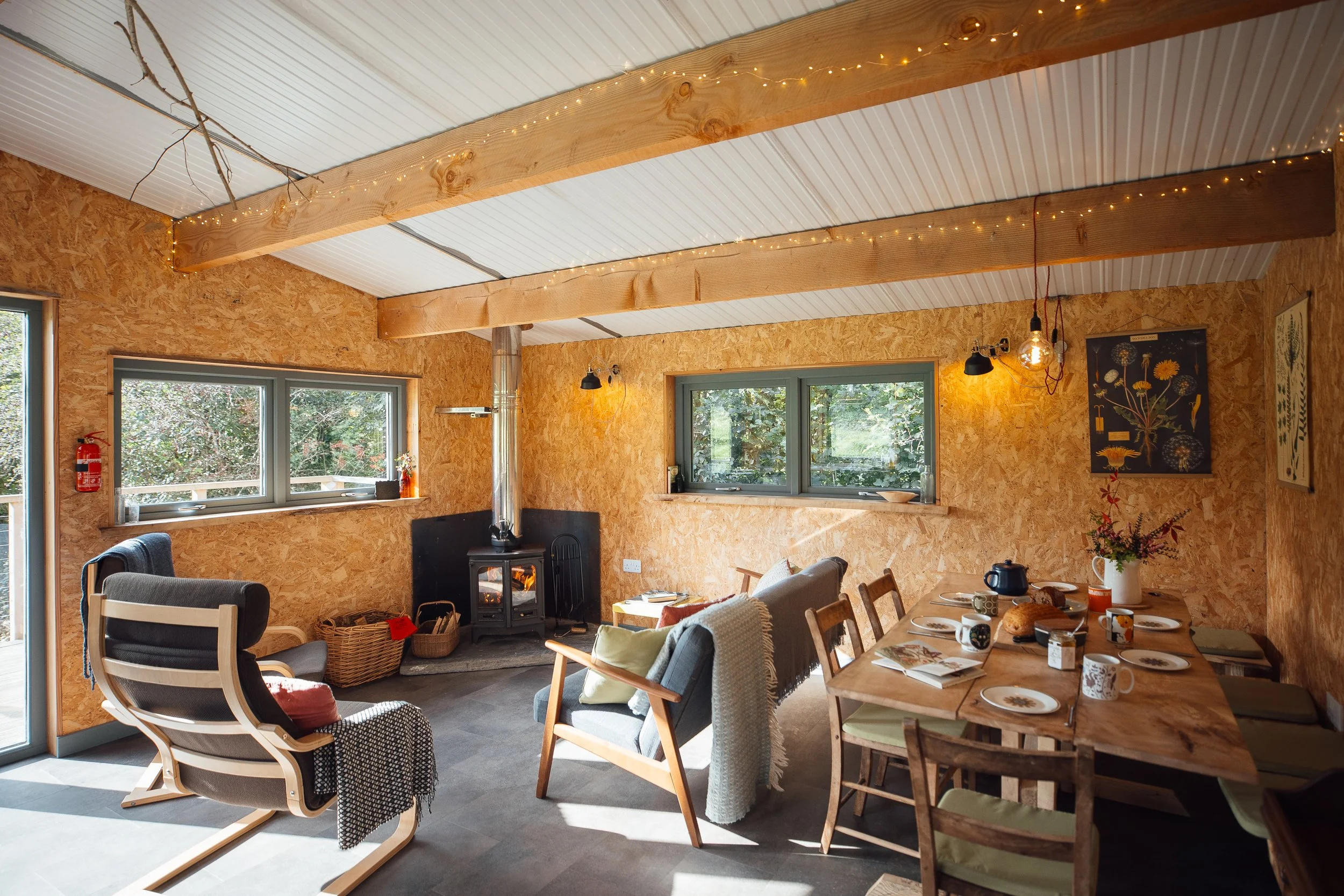 Inside Ty Mawr, the living area with period furniture in greens and greys grouped around a log burner; two picture windows with green views.