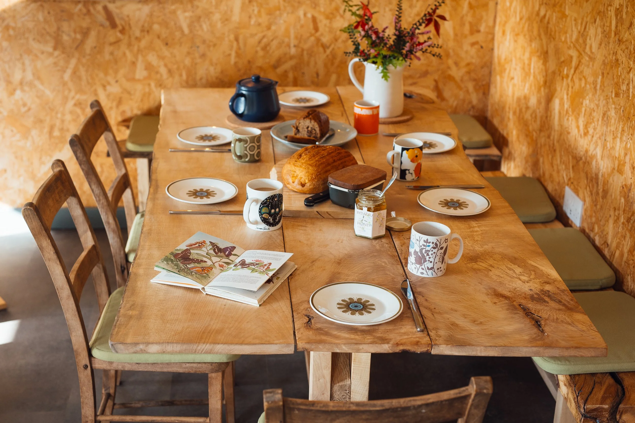View of table set for lunch at Ty Mawr, from long end of table, red flowers in a white jug. Tables seats 8-10 on chairs and stools and built in bench.
