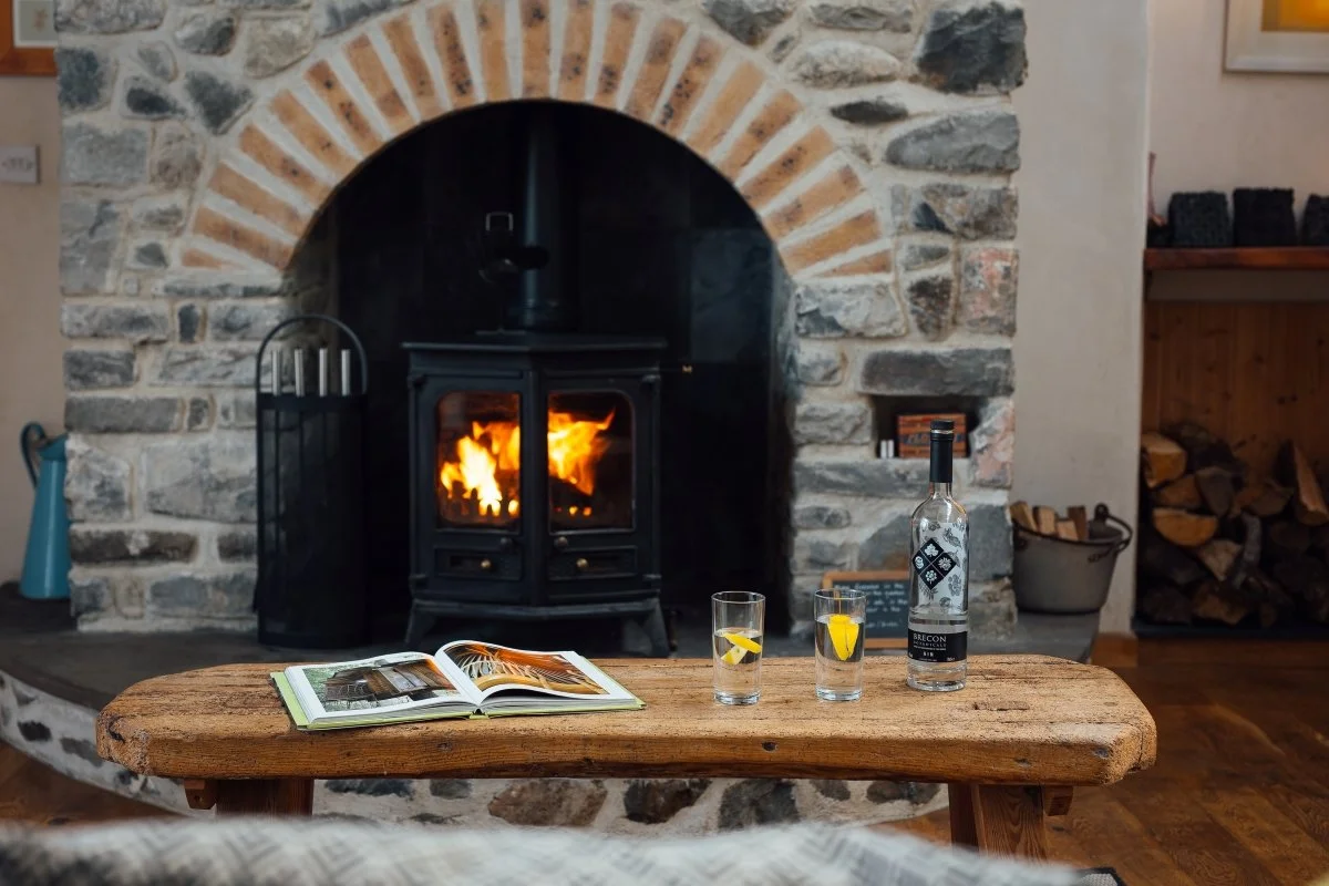 Fireplace at Llwymnbwch in stone with brick arch detail, glowing flames in black log burner. Rustic timber coffee table in foreground with picture book, bottle and two glasses.