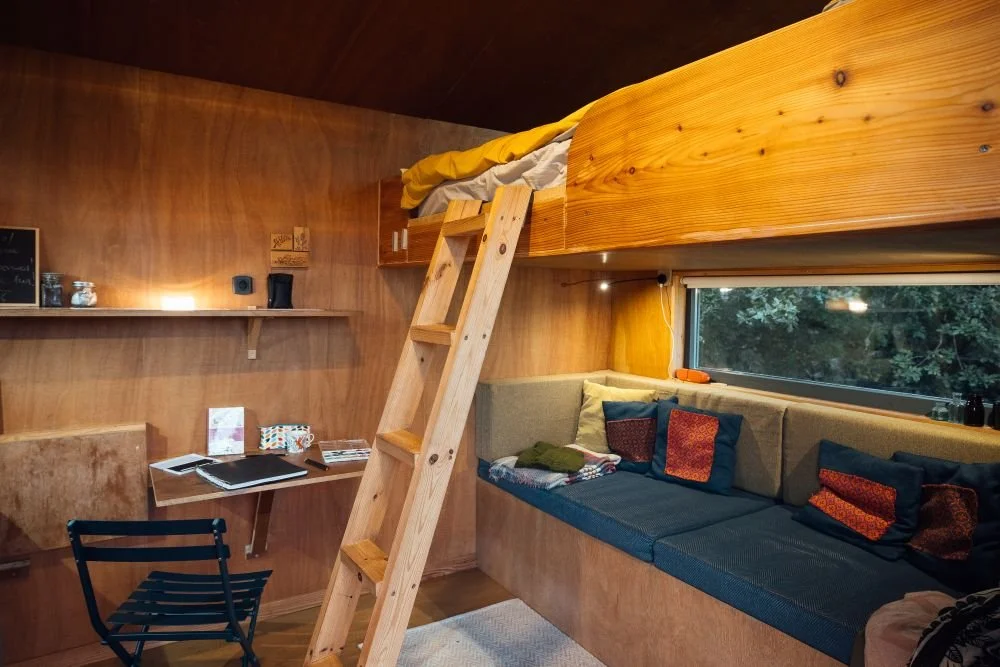 Inside Derwen, cosy lighting, showing mezzanine bed above with ladder access and blue sofa pullout bed below,blue metal chair and small table set up for writing.