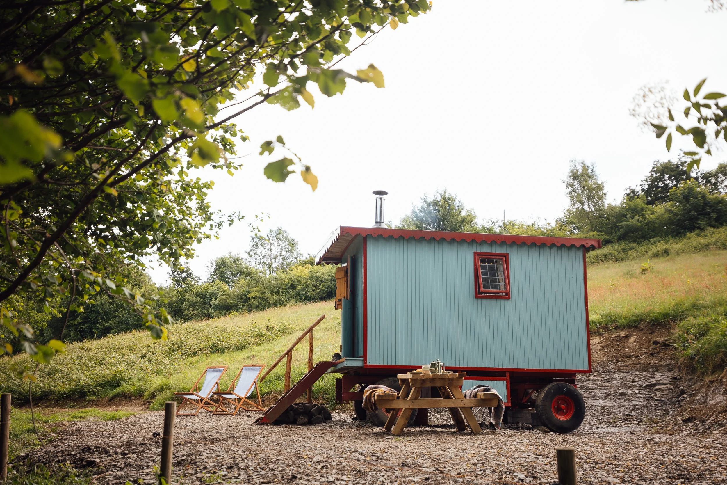 Small blue and red tiny wagon with a chimney, set on a grassy hillside landscape with trees, alongside two wooden deck chairs and a picnic table with benches on gravel ground.