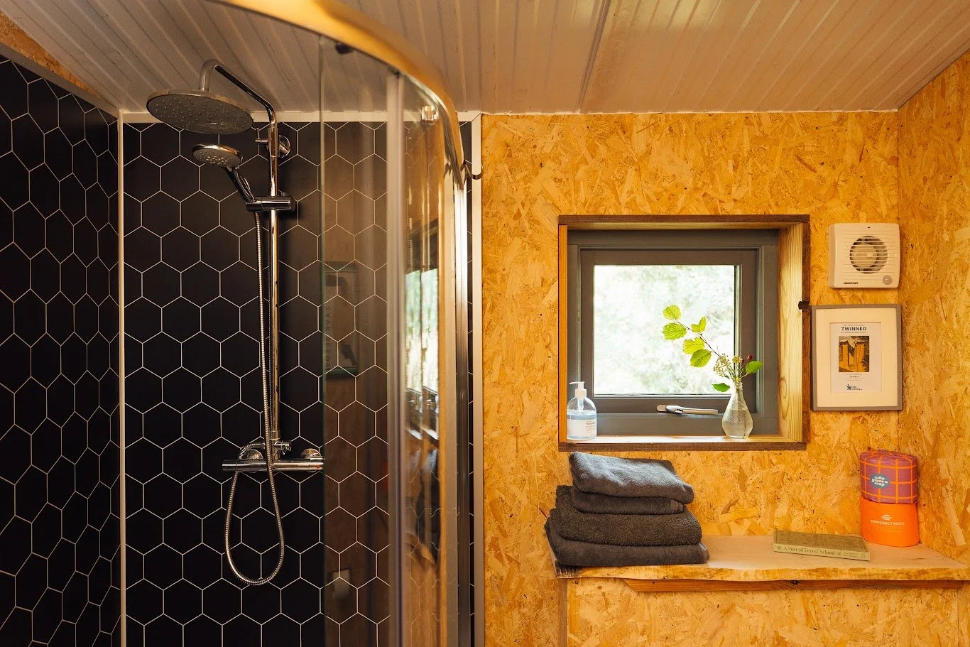 Bathroom at TY Mawr with black honeycomb detail splashback to the rainfall shower, timber walls, grey towels on the side with art and picture windows.
