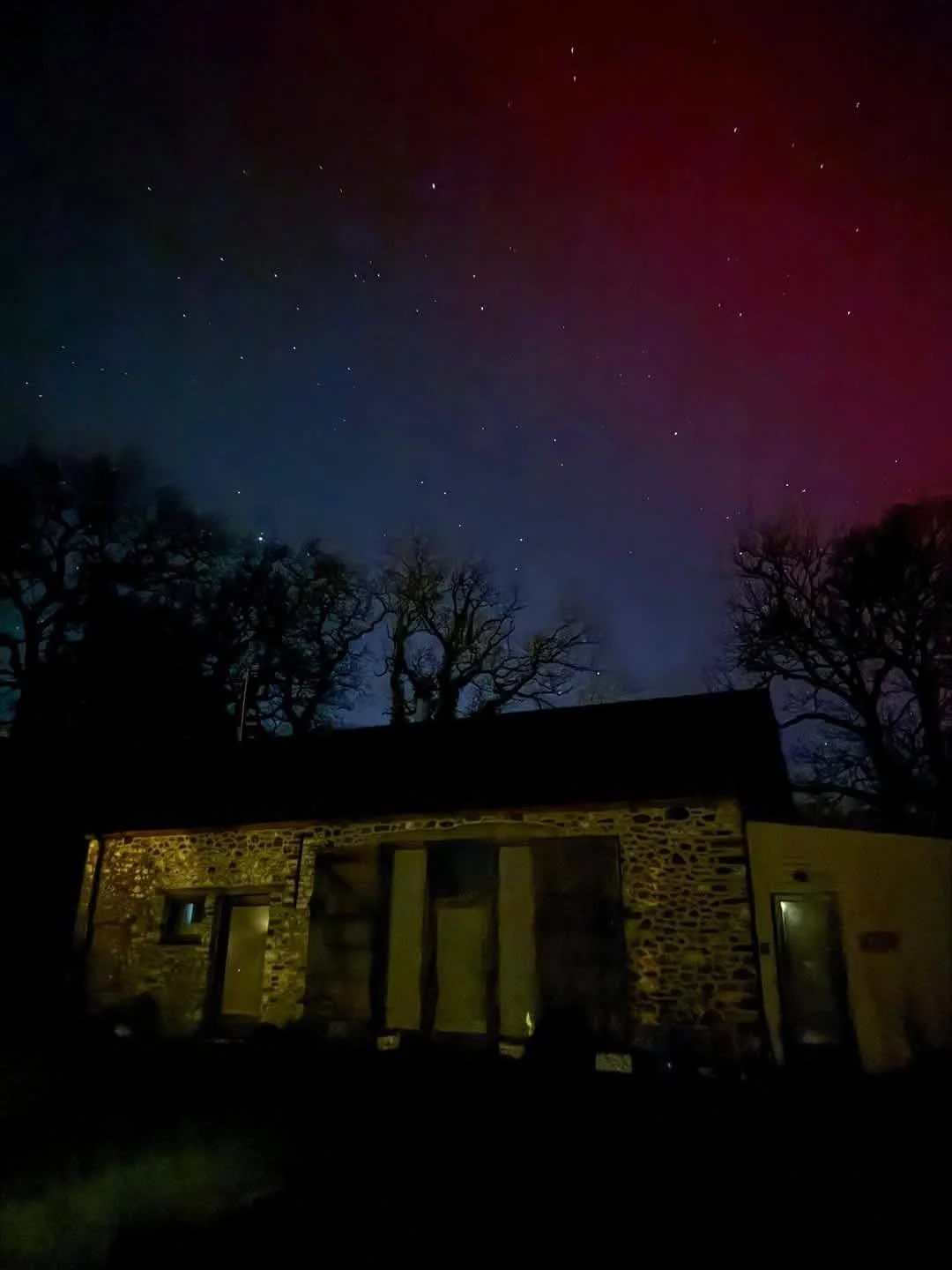Night sky with stars and colorful aurora above a stone house with trees in the background.