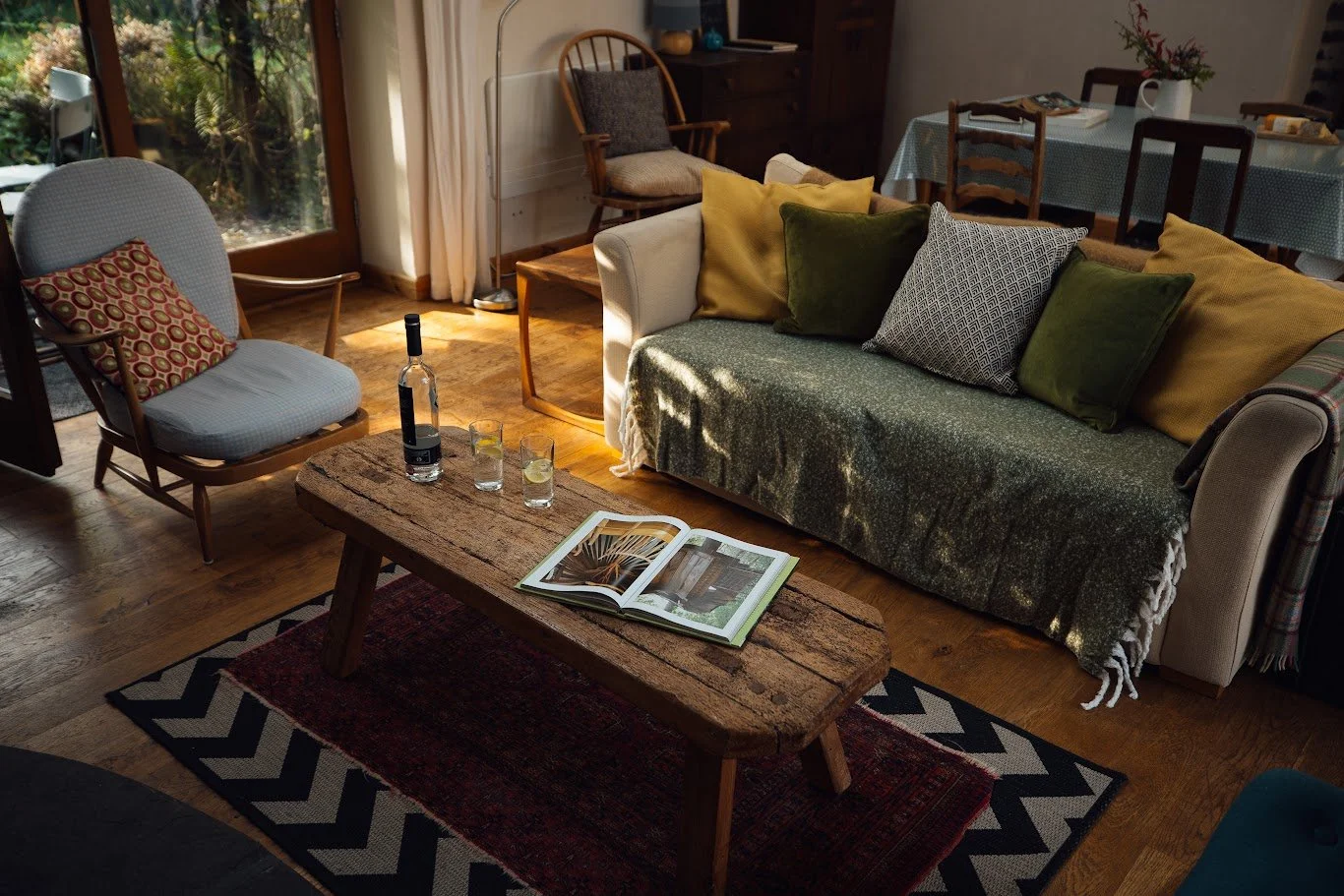 Living room chair and sofa detail at Llwynbwch Barn with handmade rustic coffee table, coffee table book, wine bottle and glasses, chair and sofa in greys and greens, patterned rug below, glimpse of french doors and garden beyond. Dappled sunlight t