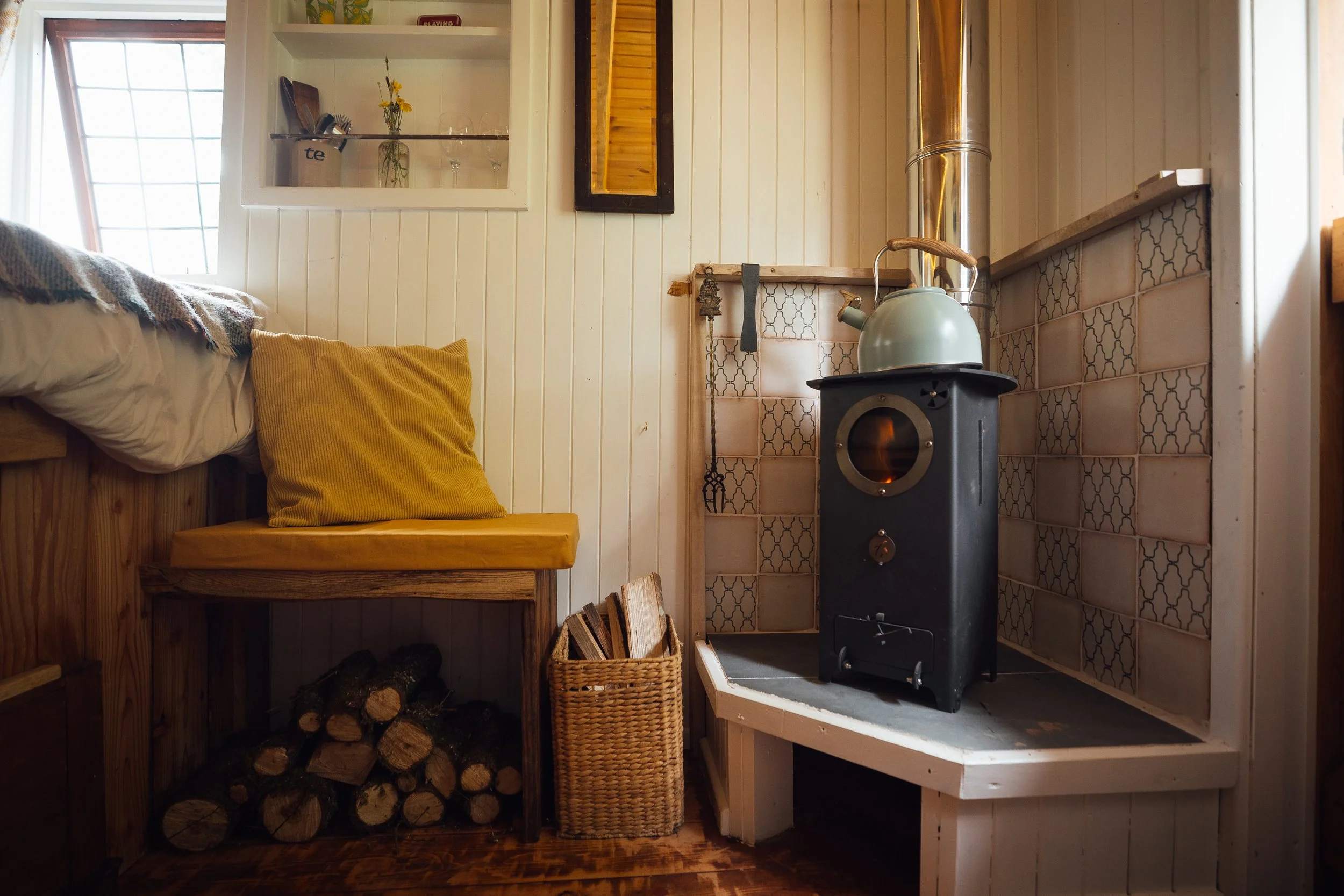 Cozy corner with a wood stove, kettle on top, a basket of firewood, a bench with yellow cushion and pillows, a window, and a shelf with a small flower vase inside a wood-paneled room.
