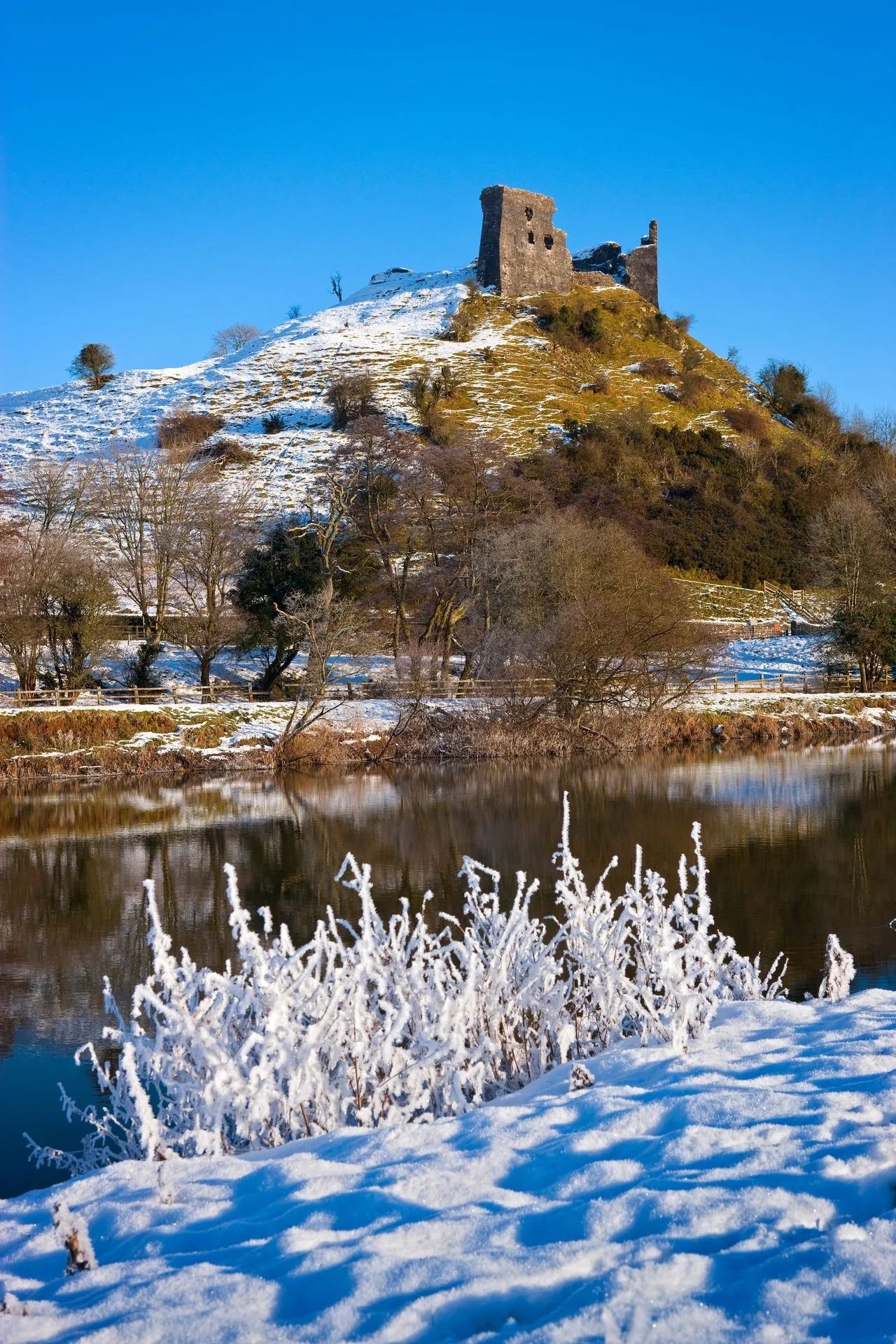 Snow-covered landscape with a river in the foreground, trees along the riverbank, and a hill with ruins of an old stone castle on top under a clear blue sky.