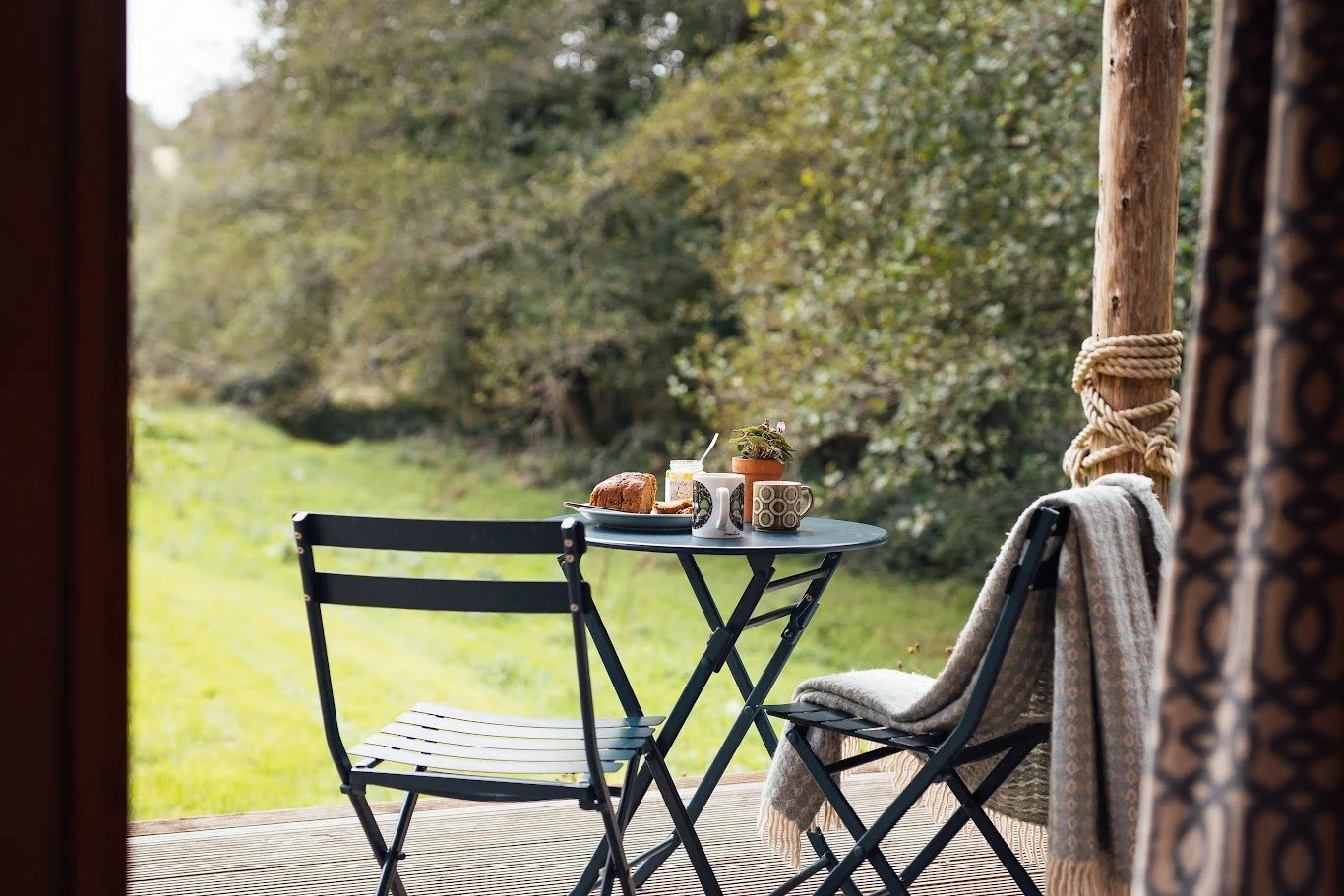 Looking out from the wooden deck of Hafan onto the grassy meadow and green hedge / trees boundary. In the foreground outdoor table & chairs laid for breakfast.