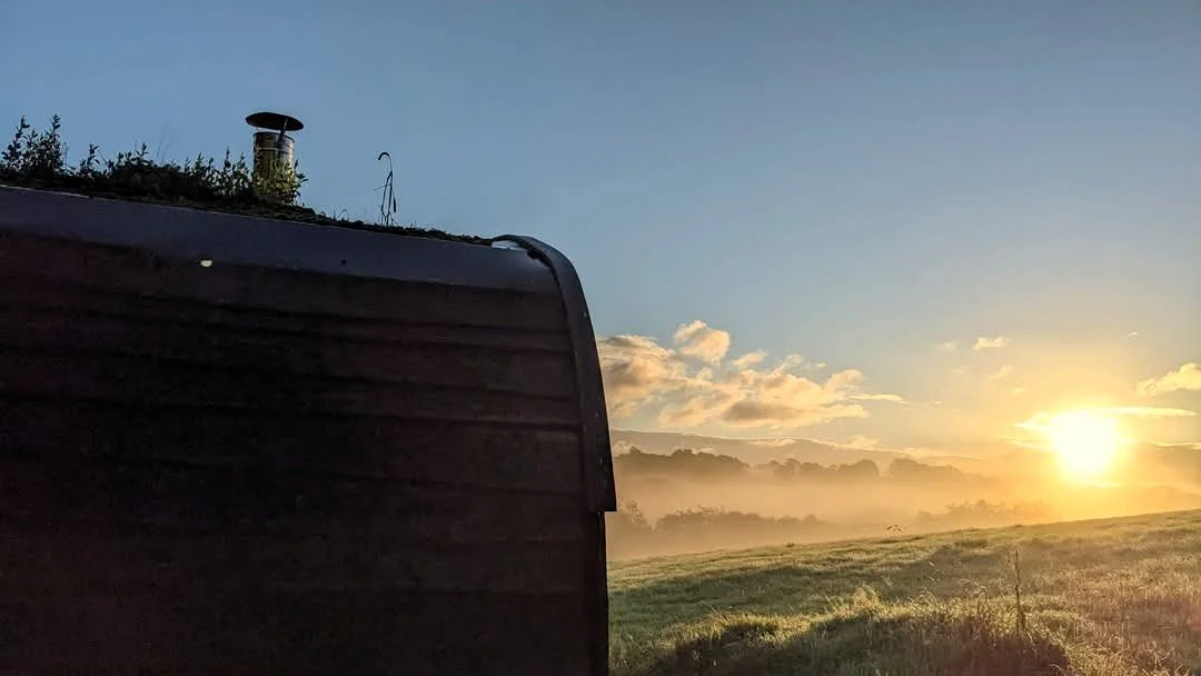 Sunrise at Derwen; the cabin to the left with its metal flu ad green roof in dark shade with low lying cloud shrouded in mist and meadow all made golden by the morning sunlight.