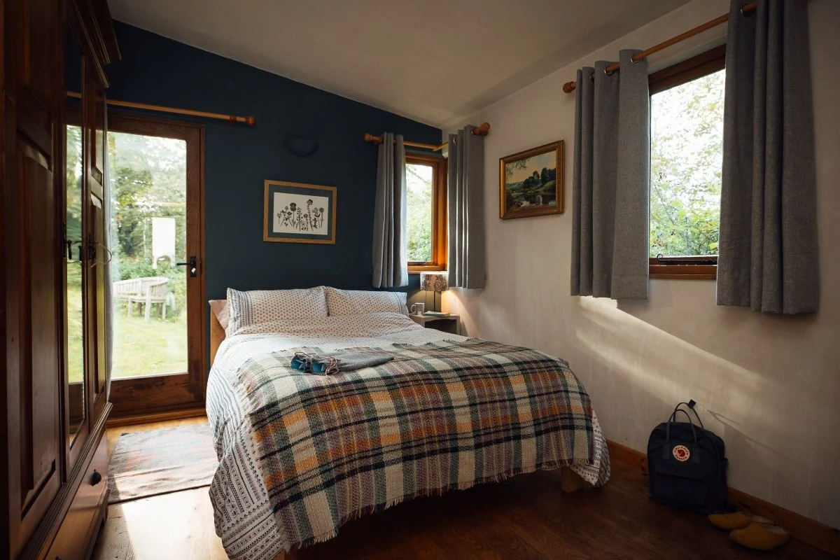 Bedroom at Llwnbwch Barn with deep blue and white walls, wooden floor and double bed with white linen and a traditional Welsh checked blanket throw. Views to garden beyond through glazed door.
