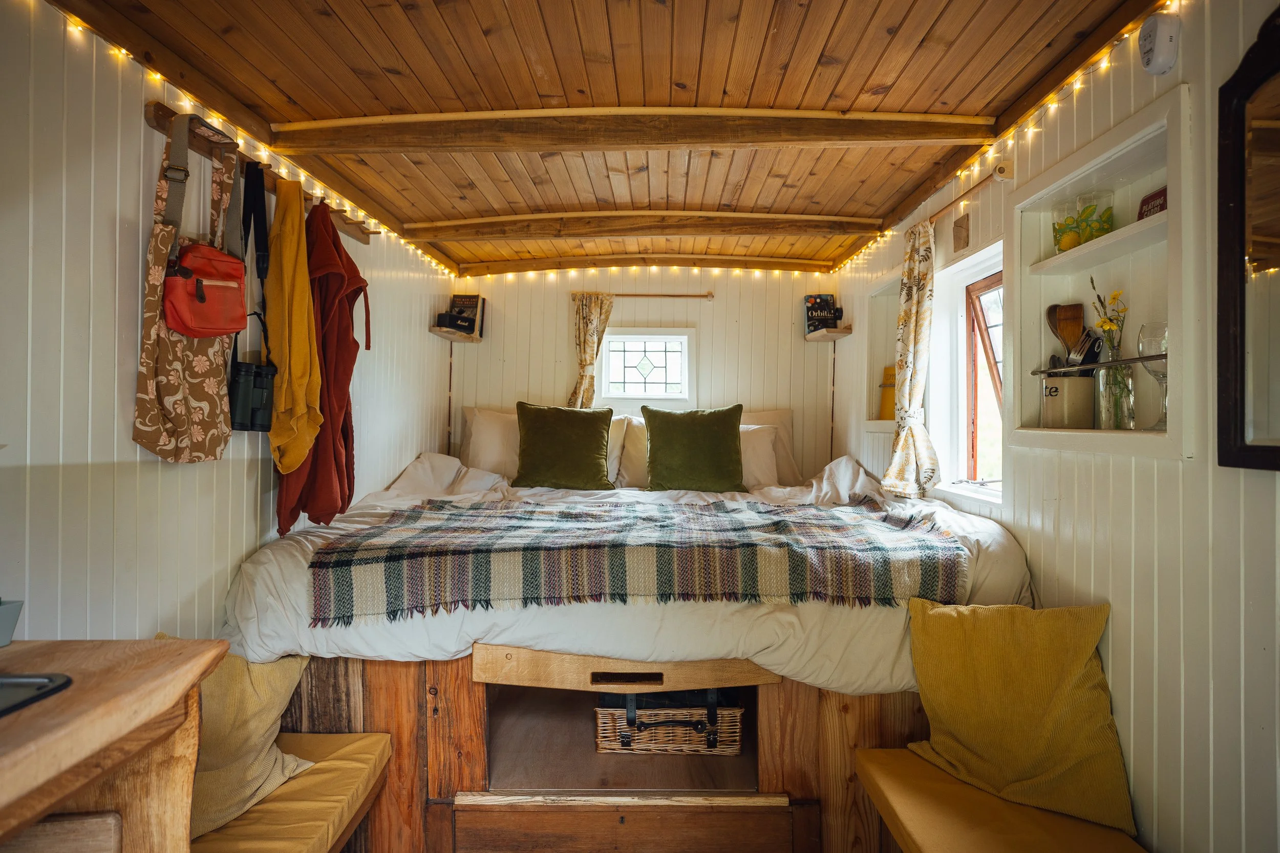 Cozy tiny bedroom with a wooden ceiling, white paneled walls, and small windows. The bed has two green pillows and a plaid blanket. There are string lights on the ceiling and a bundle of shelves and hooks with personal items on the wall.