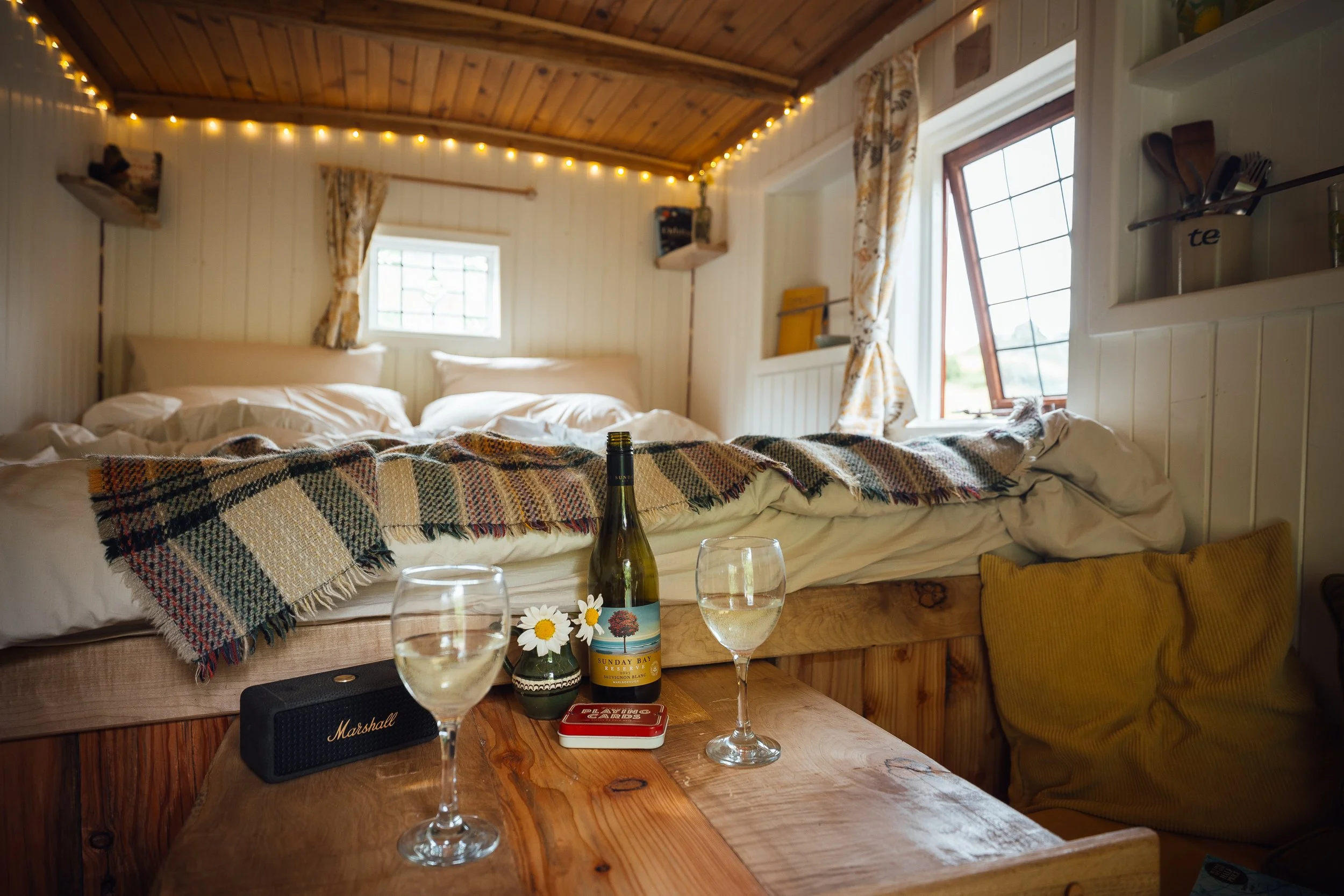 Cozy bedroom with a bed on a wooden platform, two glasses of wine on a table, a bottle of wine, and a small flower arrangement. The room features wood-paneled walls, a small window with curtains, and fairy lights along the ceiling.