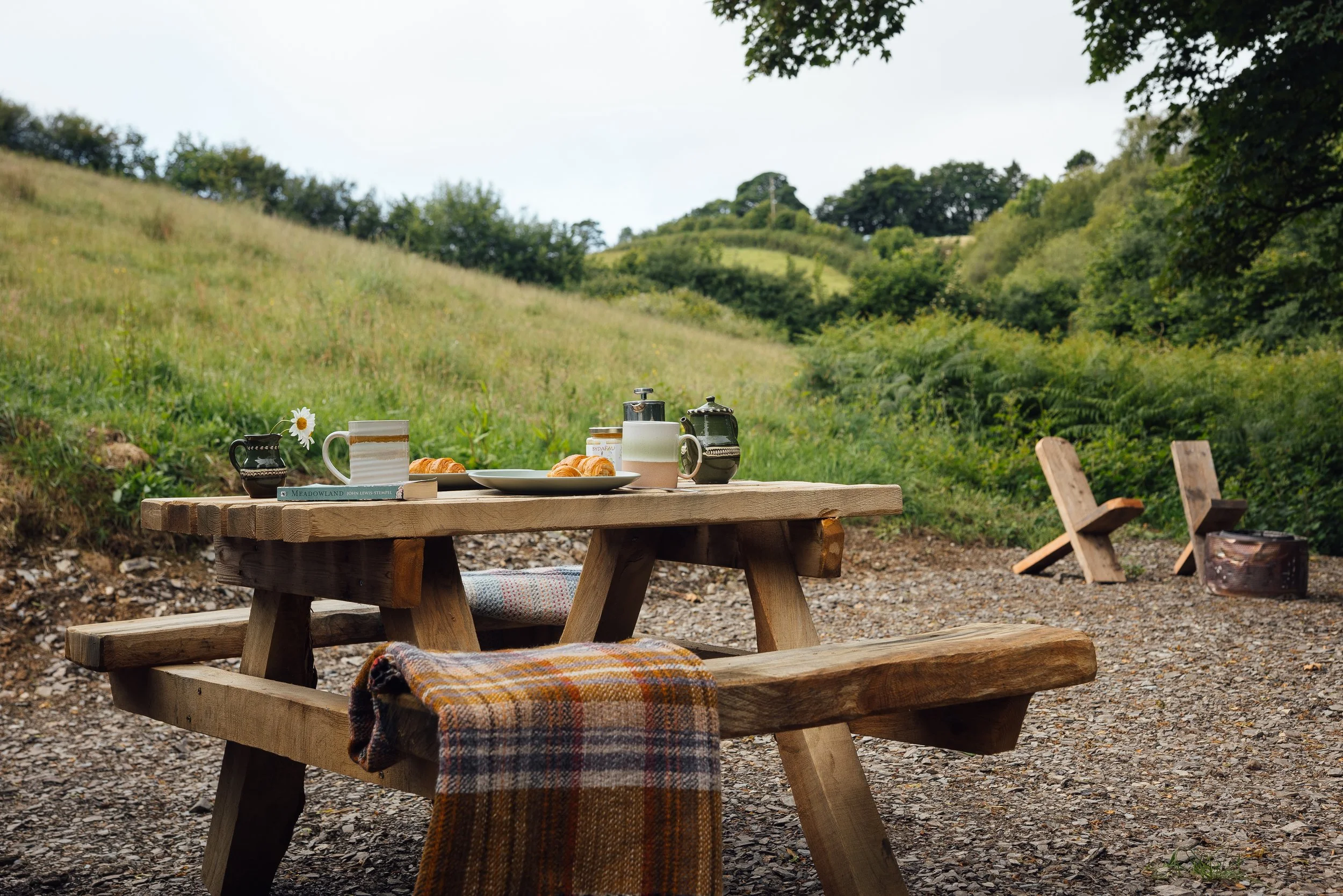 A rustic outdoor picnic table set for breakfast in a natural, green hillside setting, with two wooden chairs, a blanket, and various breakfast items.
