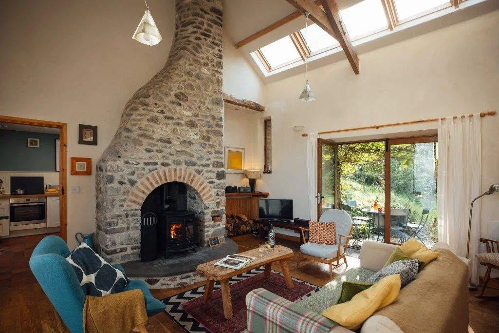 Living room at Llwynbwch Barn with wooden floor and mid century furniture in blue, grey, mustard with blankets and cushions, curved floor to ceiling stone fireplace with log burner and glowing orange flames. View to garden through french doors.