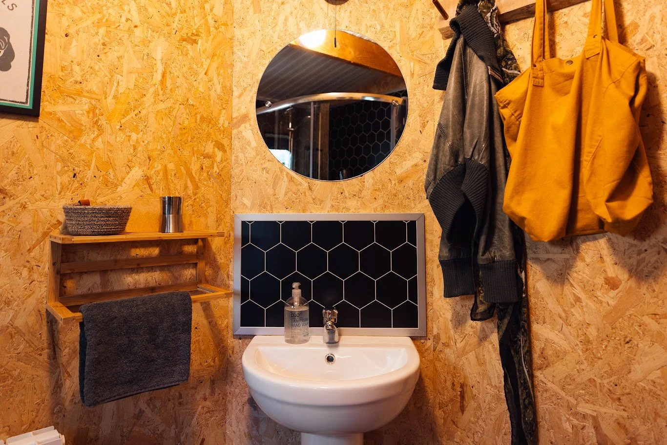 Bathroom detail in Ty Mawr focussed around the sink with black honeycomb splashback, round mirror above, wooden finish walls, a wooden towel rail and mustard yellow bag hanging from hooks.