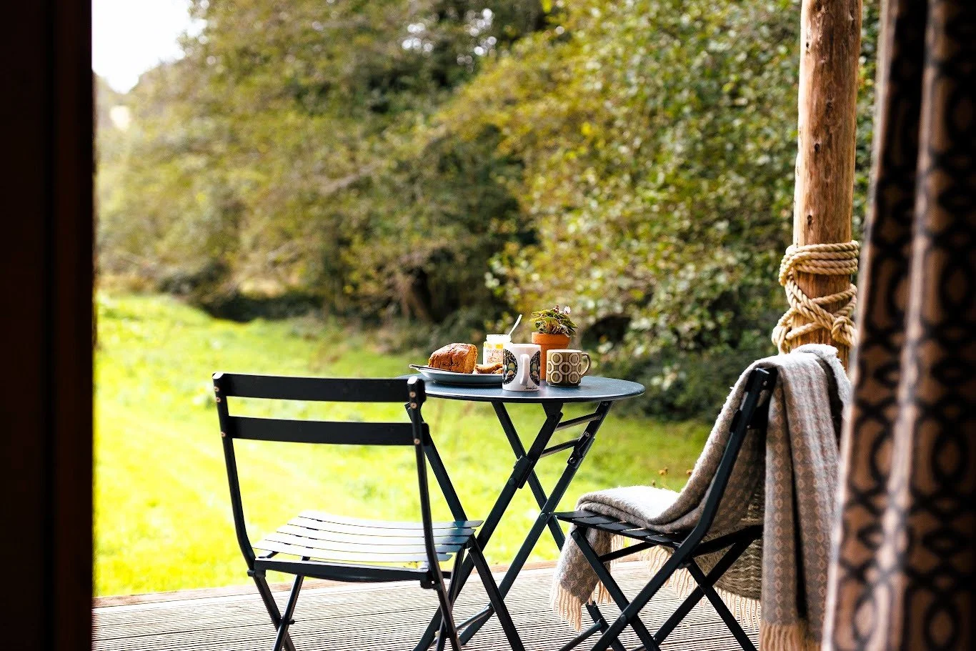 Looking out from the wooden deck of Hafan onto the grassy meadow and green hedge / trees boundary. In the foreground outdoor table & chairs laid for breakfast.