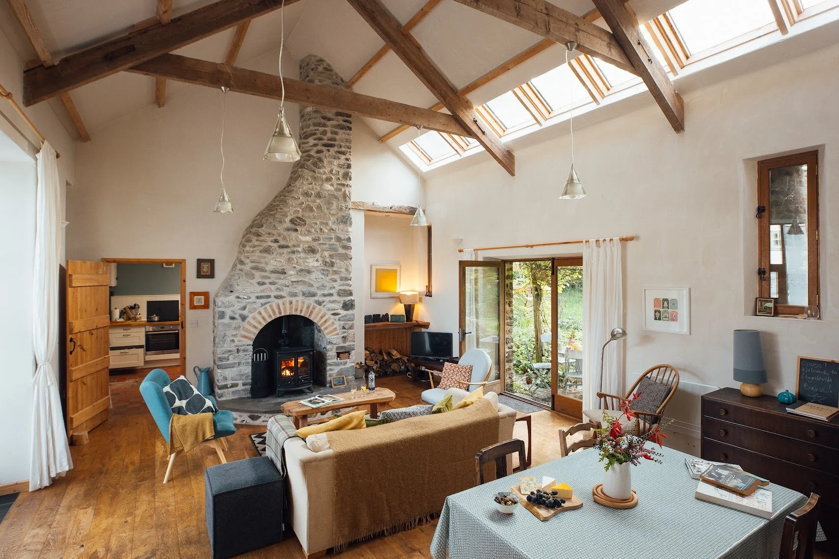Living room with a large stone fireplace, wooden ceiling beams, skylights, and a sliding glass door leading outdoors. Contains a beige sofa, several chairs, a coffee table, and a dining table with a vase of flowers.