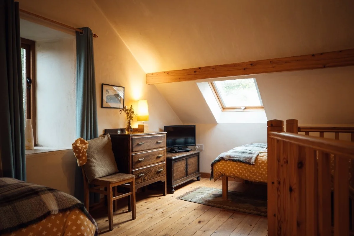 Twin bedroom at Llwynbwch with skylight, cream walls with warm timber beams and traditional furniture - timber chair, chest of drawers, two single beds with Welsh checked blanket drapes.