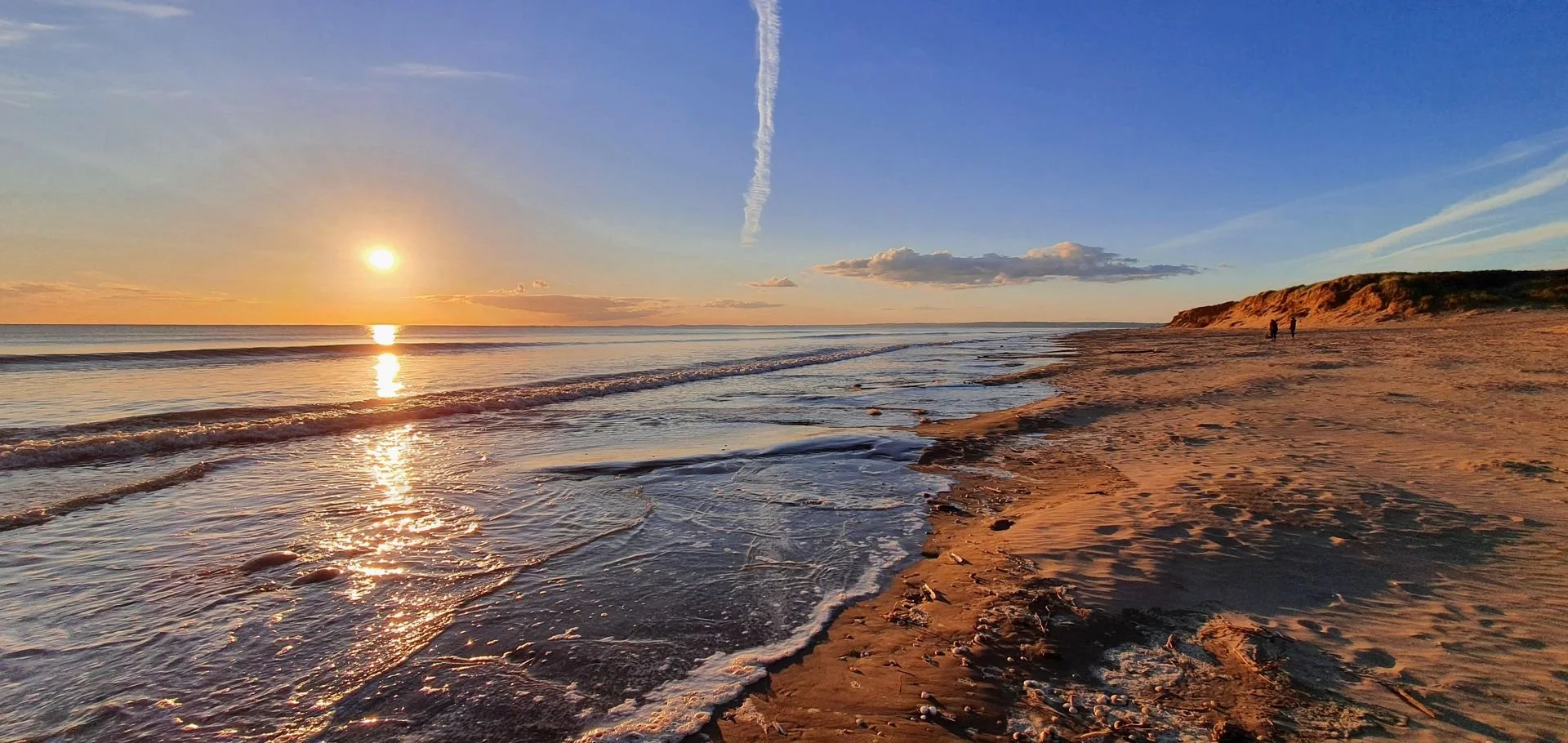 Sunset over the sea at a sandy beach with a few people walking, waves gently lapping the shore, and a hill in the background.