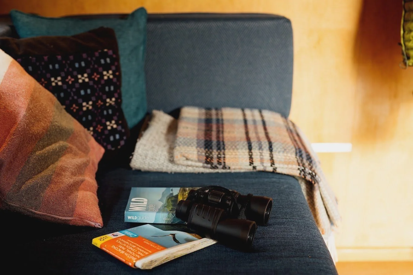 A blue couch with colourful cushions , a welsh blanket, binoculars, a guidebook, and a map on it, in a cozy room with yellow walls.