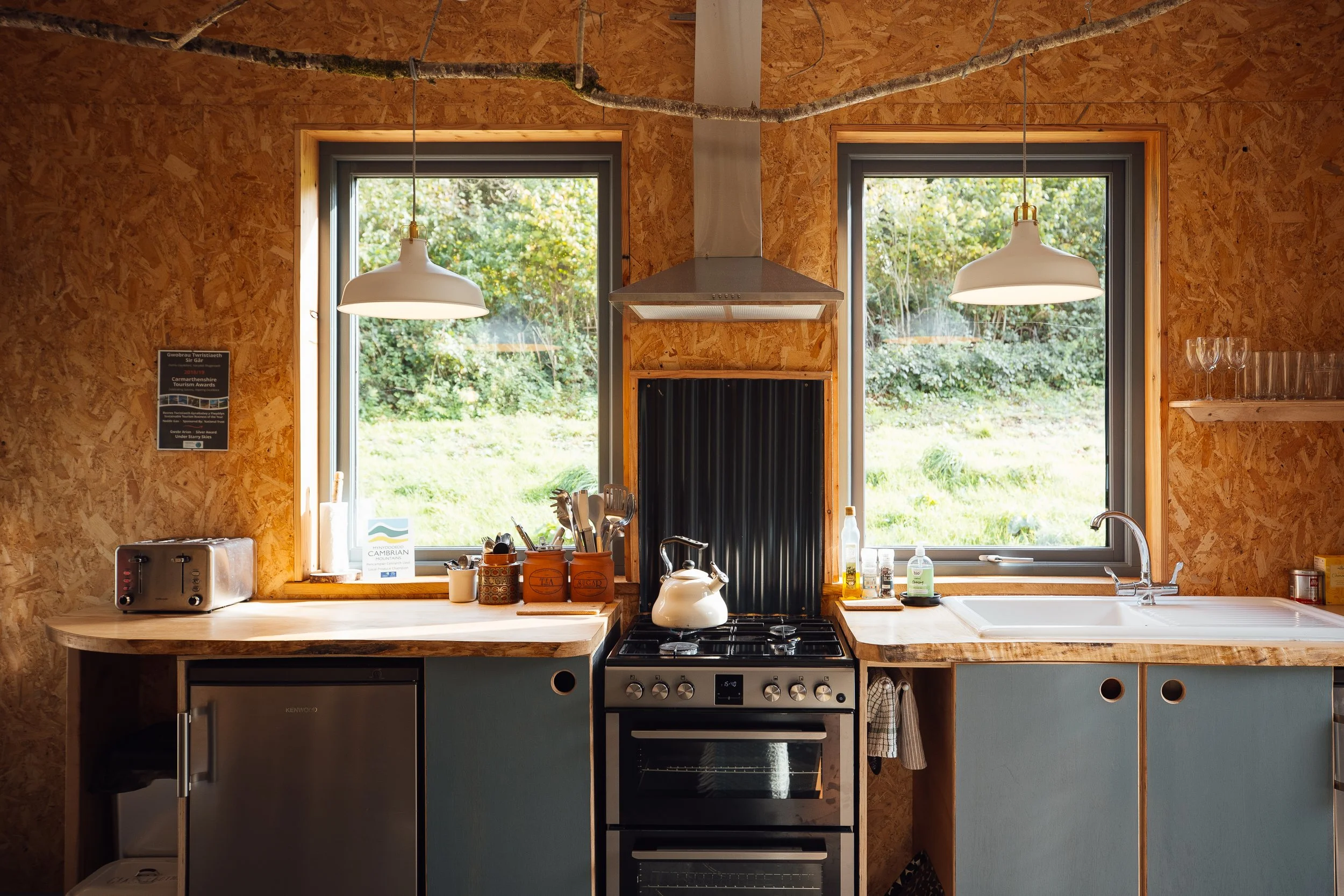 The kitchen at Ty Mawr, cooker in the centre, blue cupboards below and focal point is two picture windows looking onto the green space beyond.