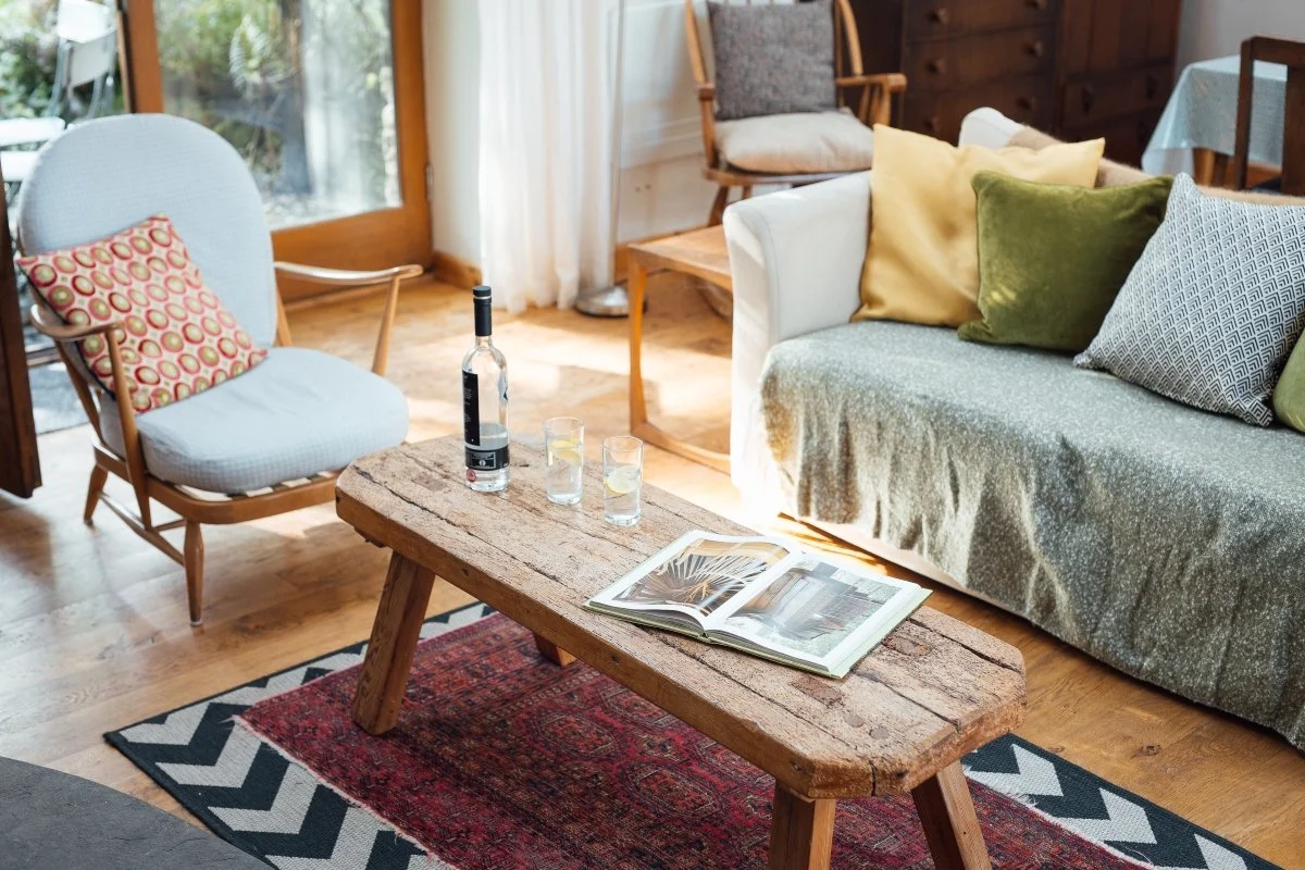 Living room chair and sofa detail at Llwynbwch Barn with handmade rustic coffee table, coffee table book, wine bottle and glasses, chair and sofa in greys and greens, patterned rug below, glimpse of french doors and garden beyond.