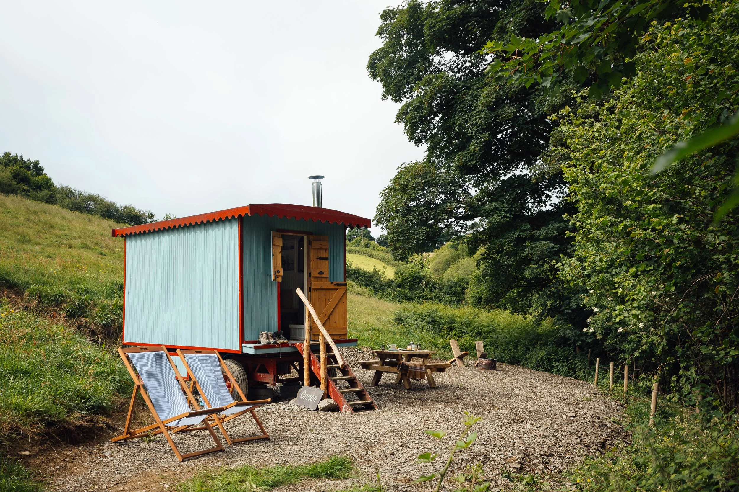 Colourful tiny wagon with a gravel path, outdoor chairs, and picnic tables in a lush green hillside setting.
