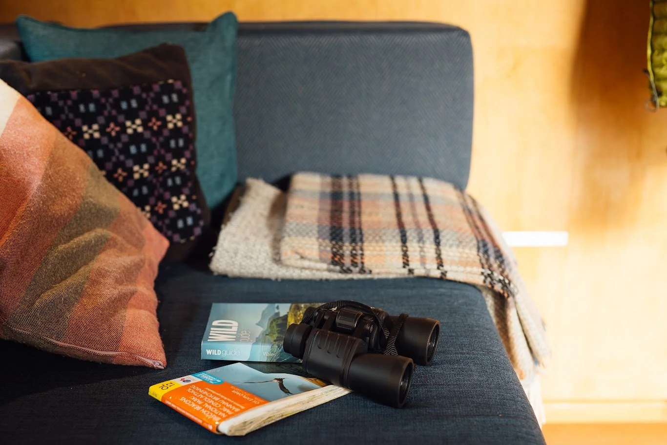 Detail of the blue sofa inside Hafan cabin with checked blanket, cushions, binoculars, map and book.