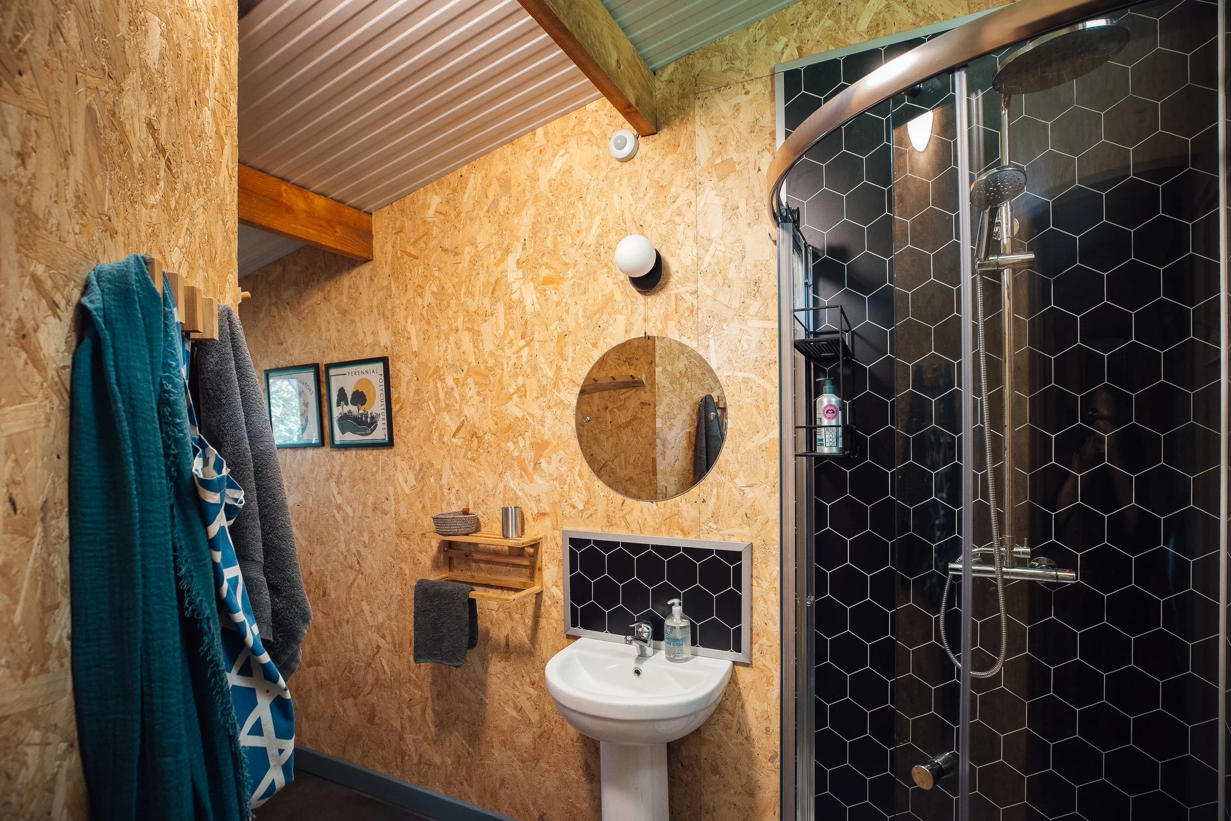 Bathroom in Ty Mawr - the sink (and shower with black honeycomb splashbacks, round mirror above, wooden finish walls, and towels hanging from wooden hooks. Corrugated roof with timber beams.