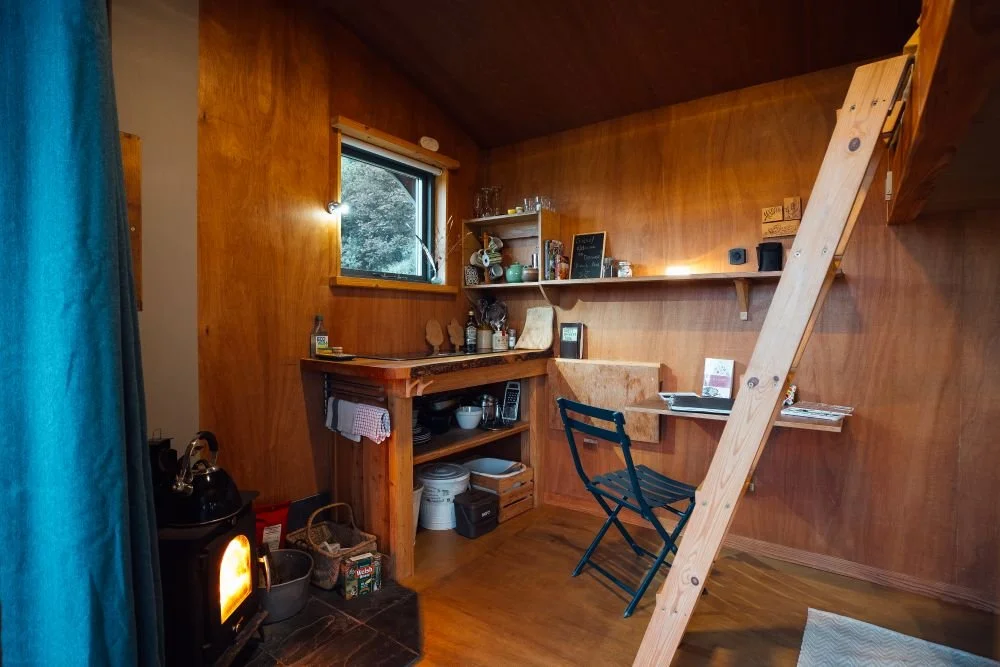Interior kitchen and dining space at Derwen Cabin with blue curtain and log burner to left and bunk bed ladder to right. Window with green view, all interior walls wooden.