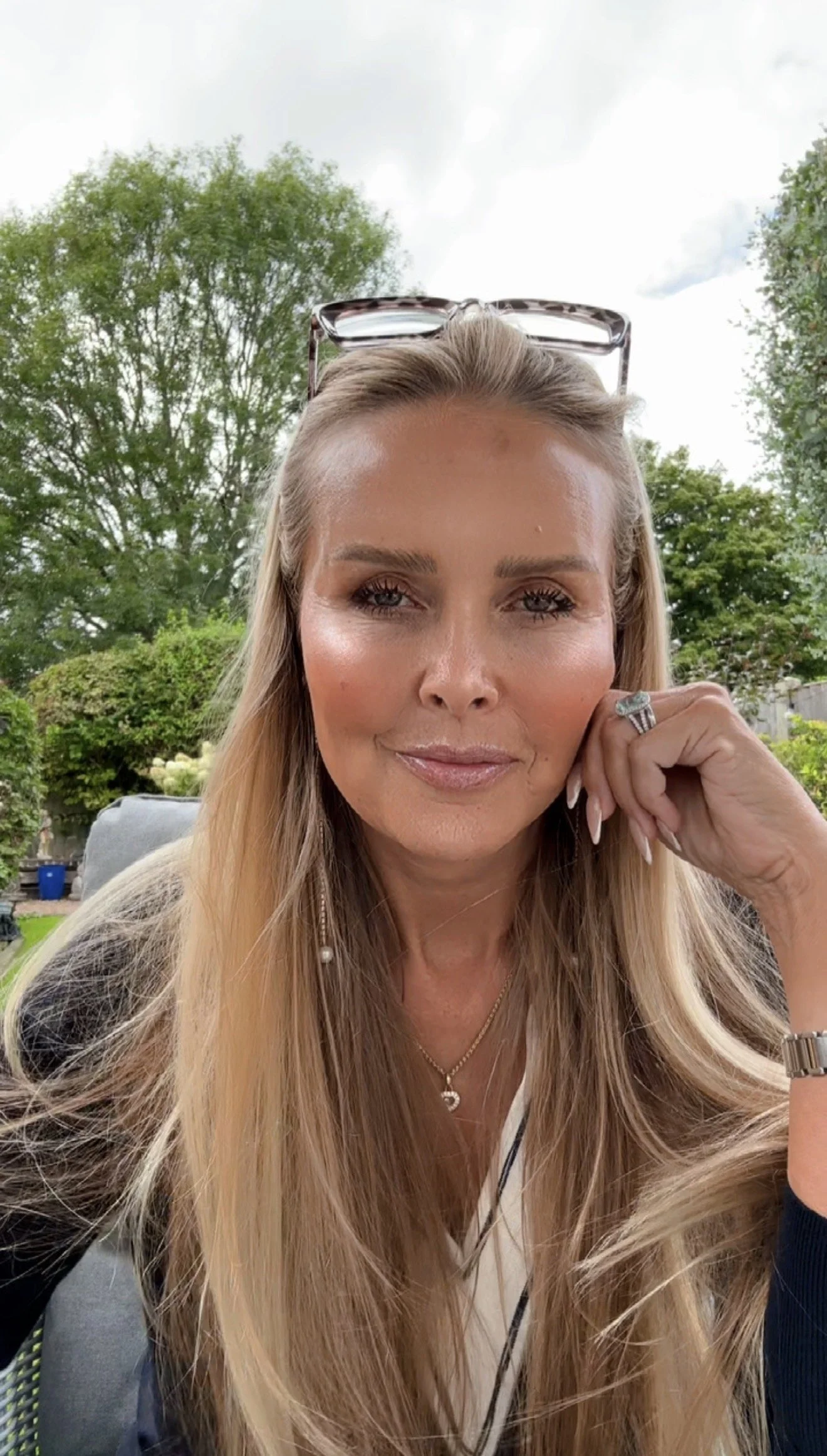 Blonde woman taking a close-up outdoor selfie, wearing glasses on her head and a large ring.