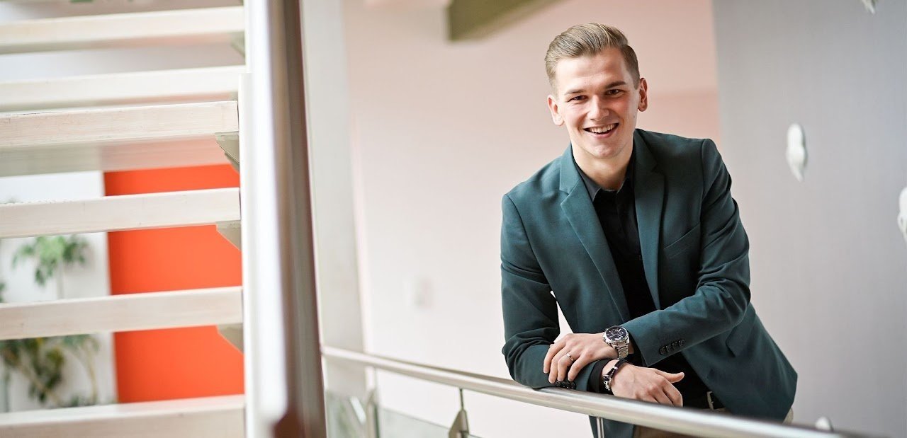 Ein lächelnder Mann in einem dunklen Anzug lehnt sich an ein Geländer in einem modernen, hellen Innenraum.