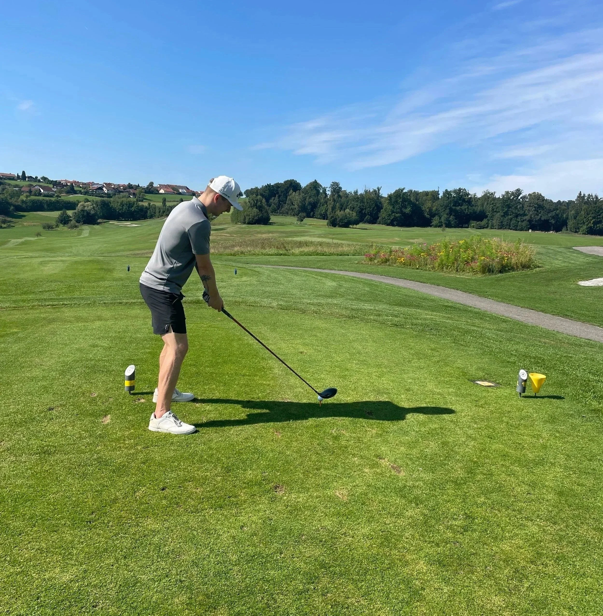 Mann bereitet sich auf einen Abschlag auf einem Golfplatz vor, umgeben von grünem Rasen, Bäumen und blauer Himmel.