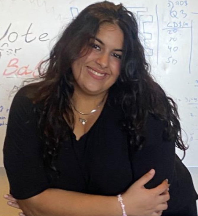 A young woman with long curly dark hair smiling and crossing her arms in front of a whiteboard with writing.